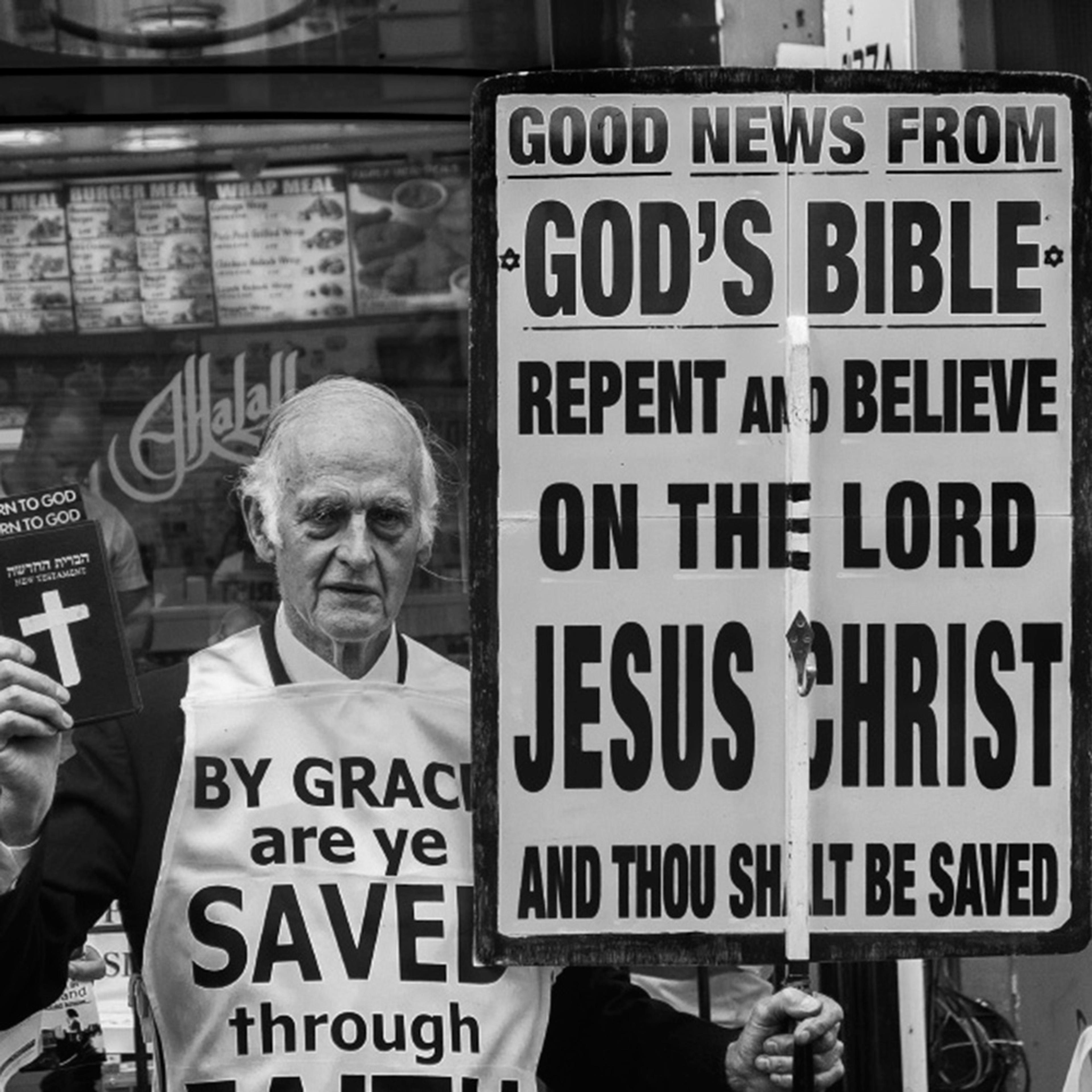 Black and white photo of an elderly man holding a Bible and a large sign with a religious message in front of a shop window.