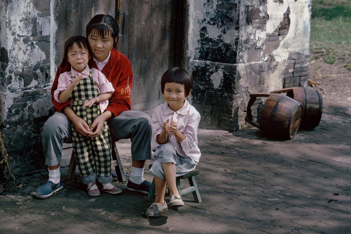 Photo of three children seated on stools by a weathered wall, with barrels in the background and one child clapping.