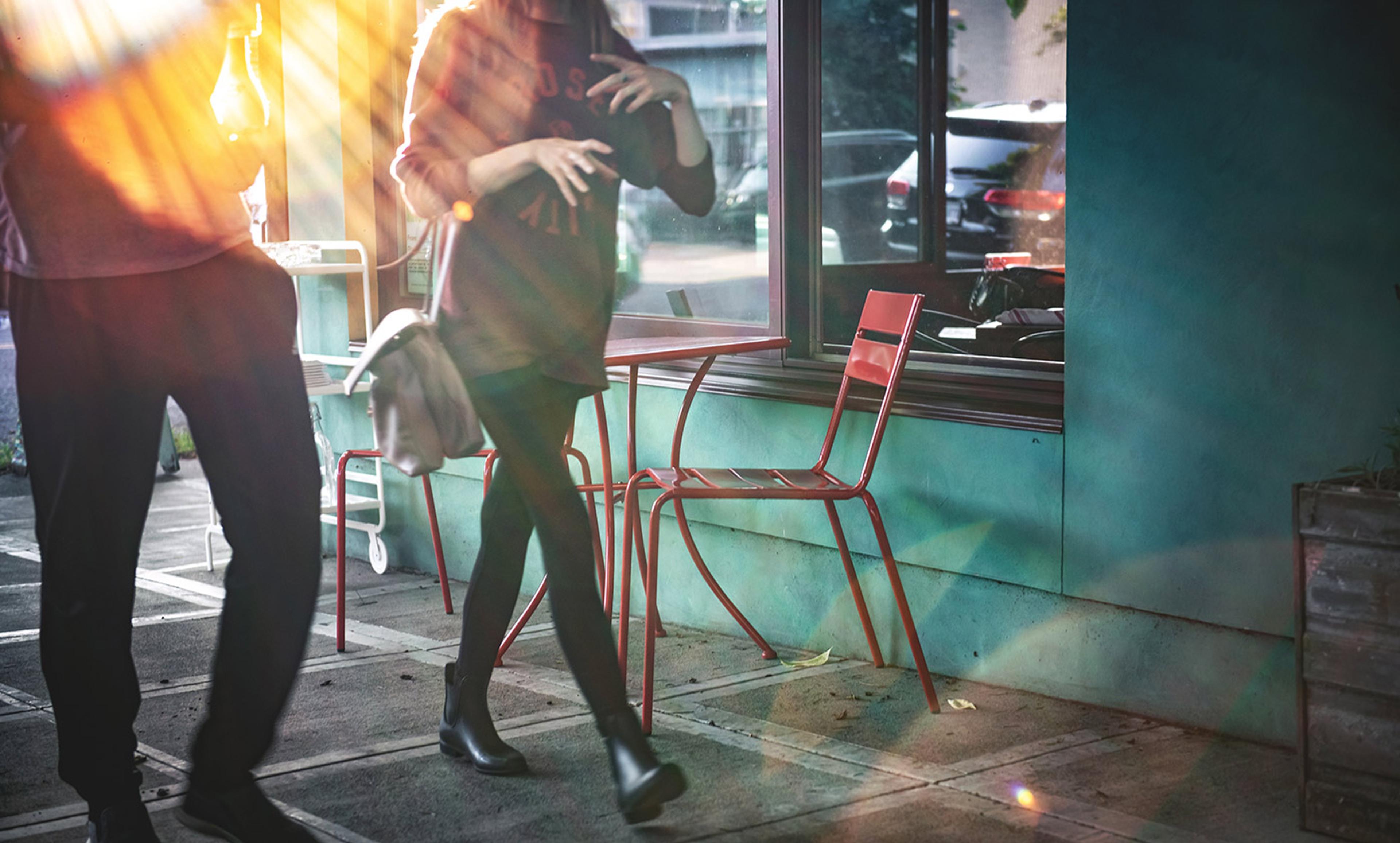 Two people walking past a sunlit cafe window, with red chairs and table outside. Sun rays create a warm glow on the scene.