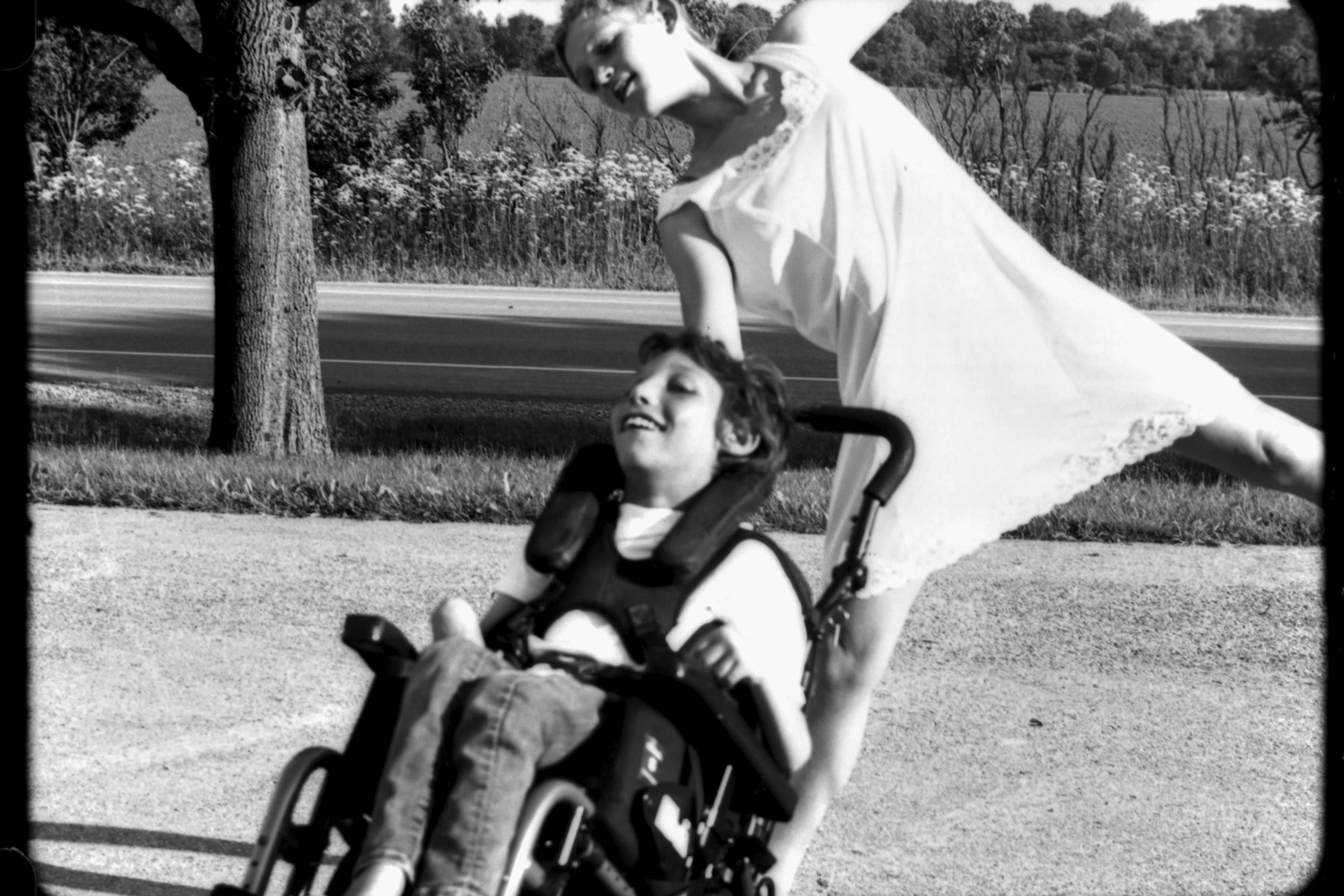 A black and white image of a girl in a white dress dancing behind her younger brother in a wheelchair, with trees and a road in the background. They are both smiling.