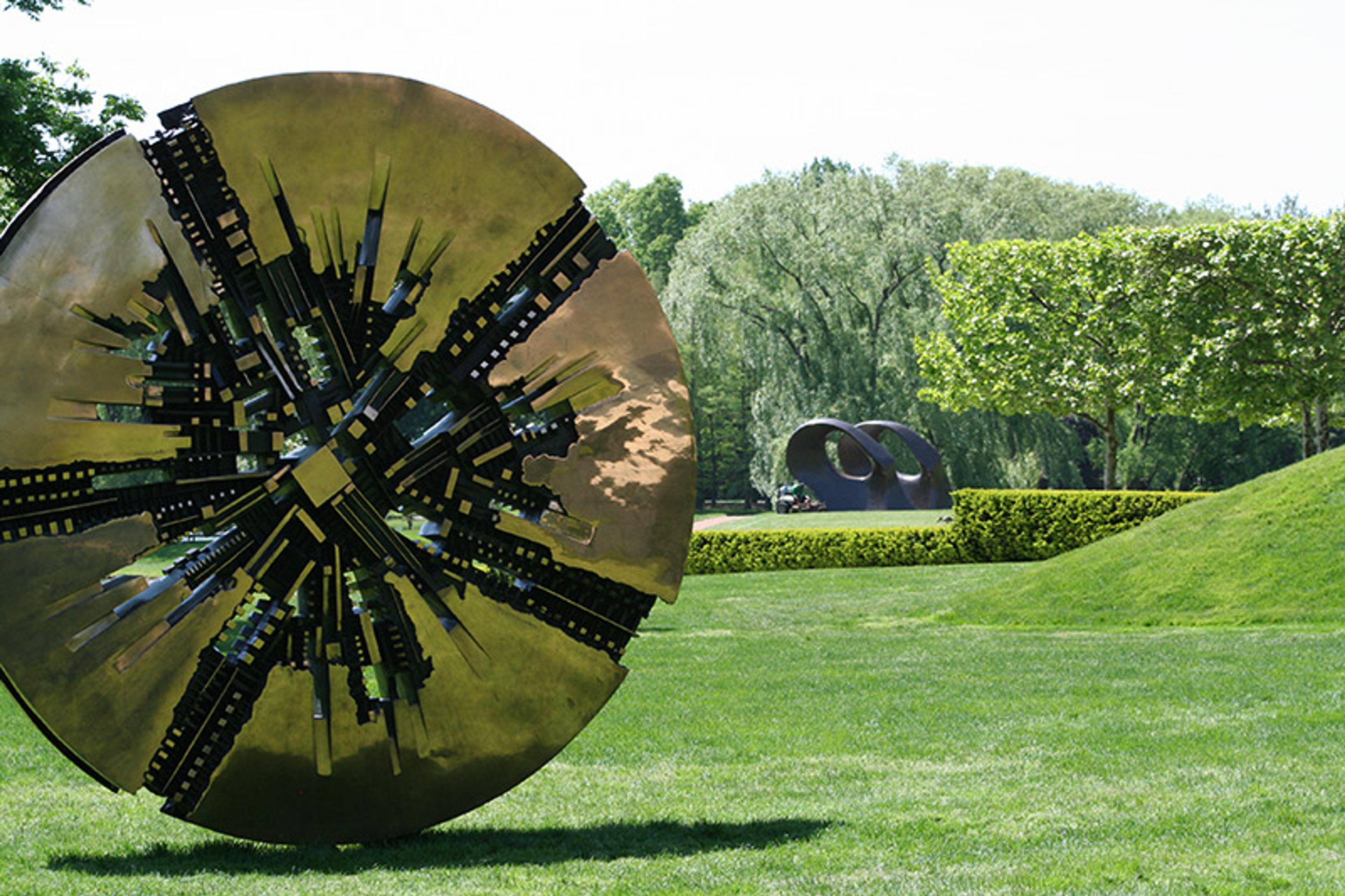 A large, abstract bronze sculpture set in a green park with trees and another sculpture in the background.