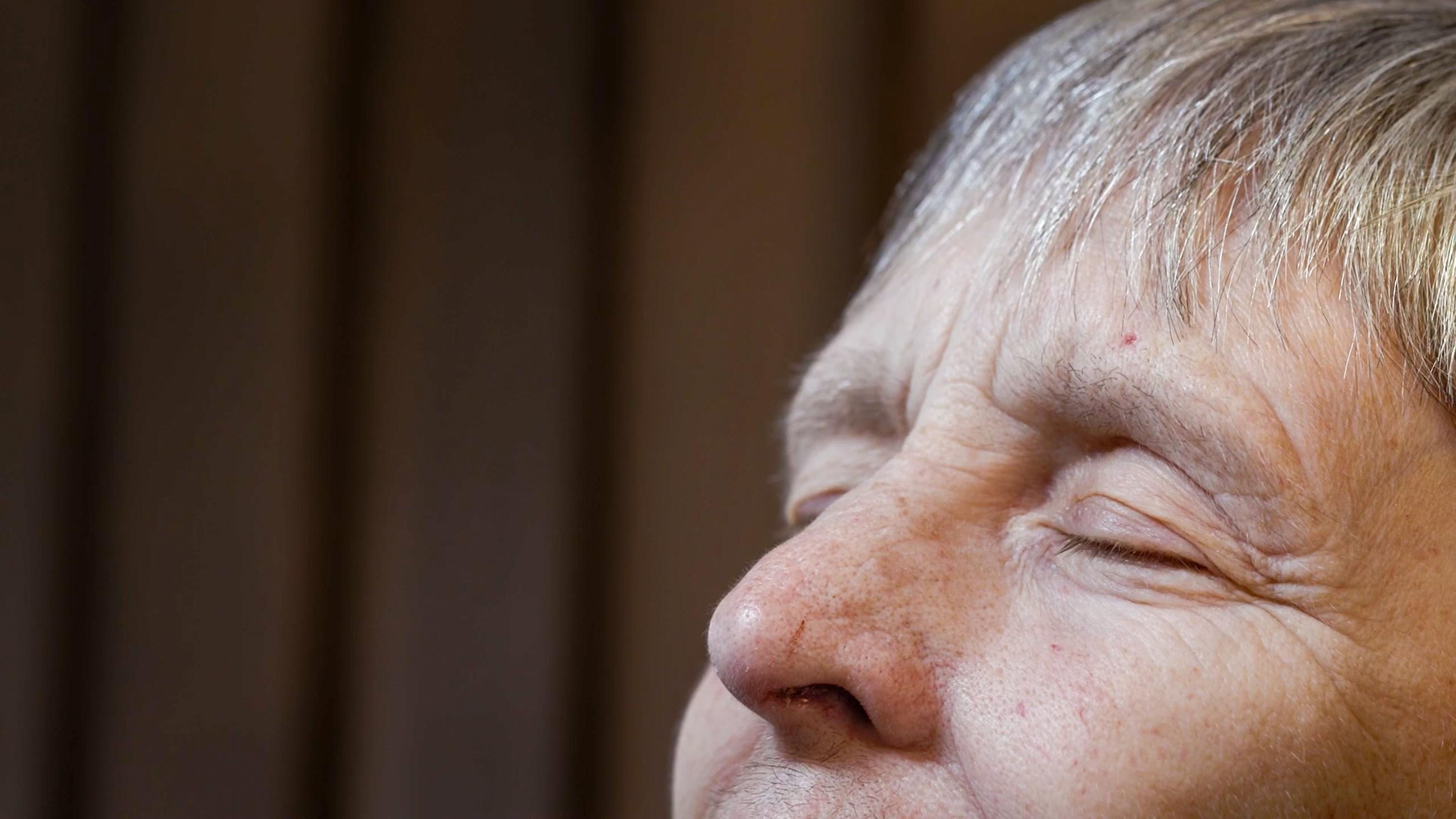 Close-up of an elderly woman’s face with eyes closed, light grey hair, and a blurred brown background. Her expression is calm.
