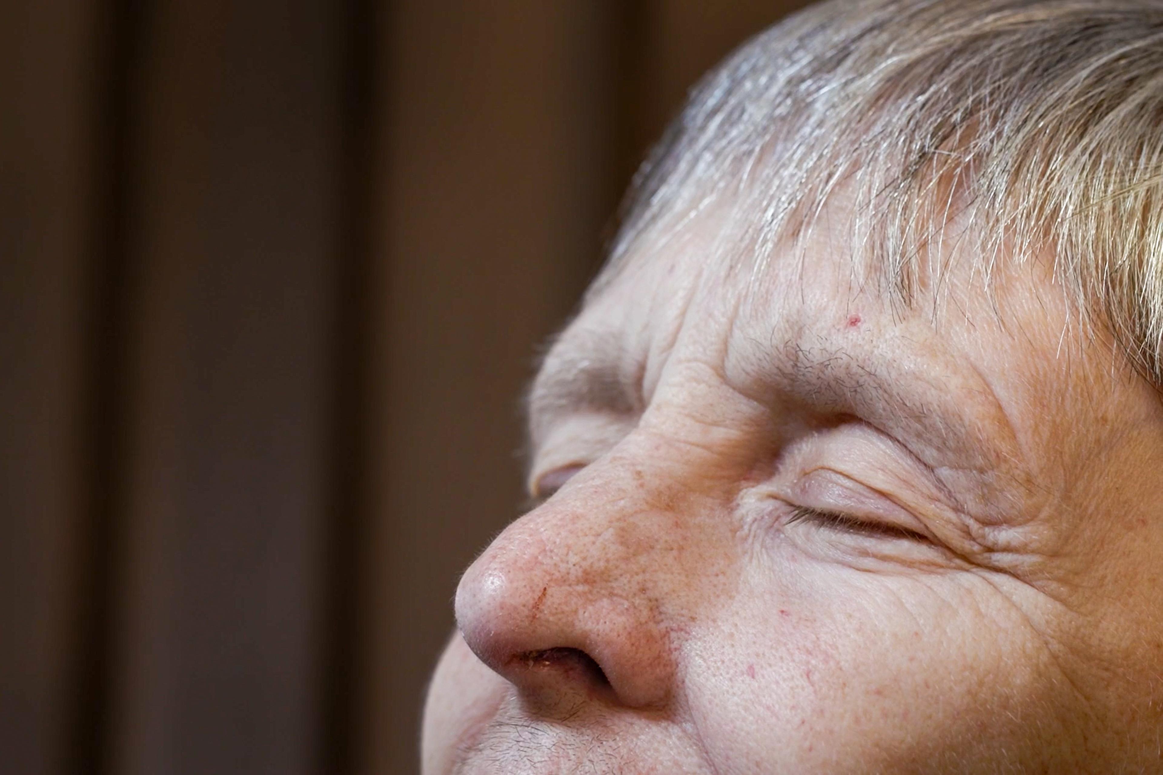 Close-up of an elderly woman’s face with eyes closed, light grey hair, and a blurred brown background. Her expression is calm.