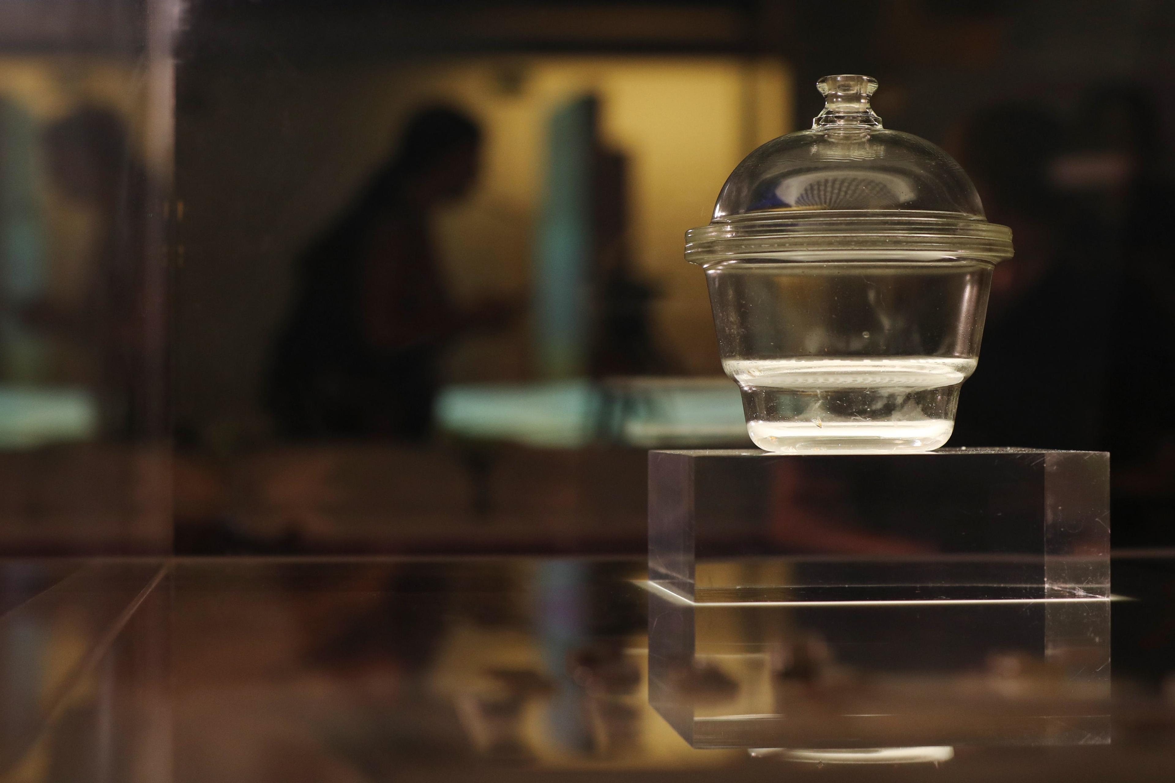 Photo of a glass jar with a lid on a pedestal in a dimly lit setting, with blurred figures in the background.