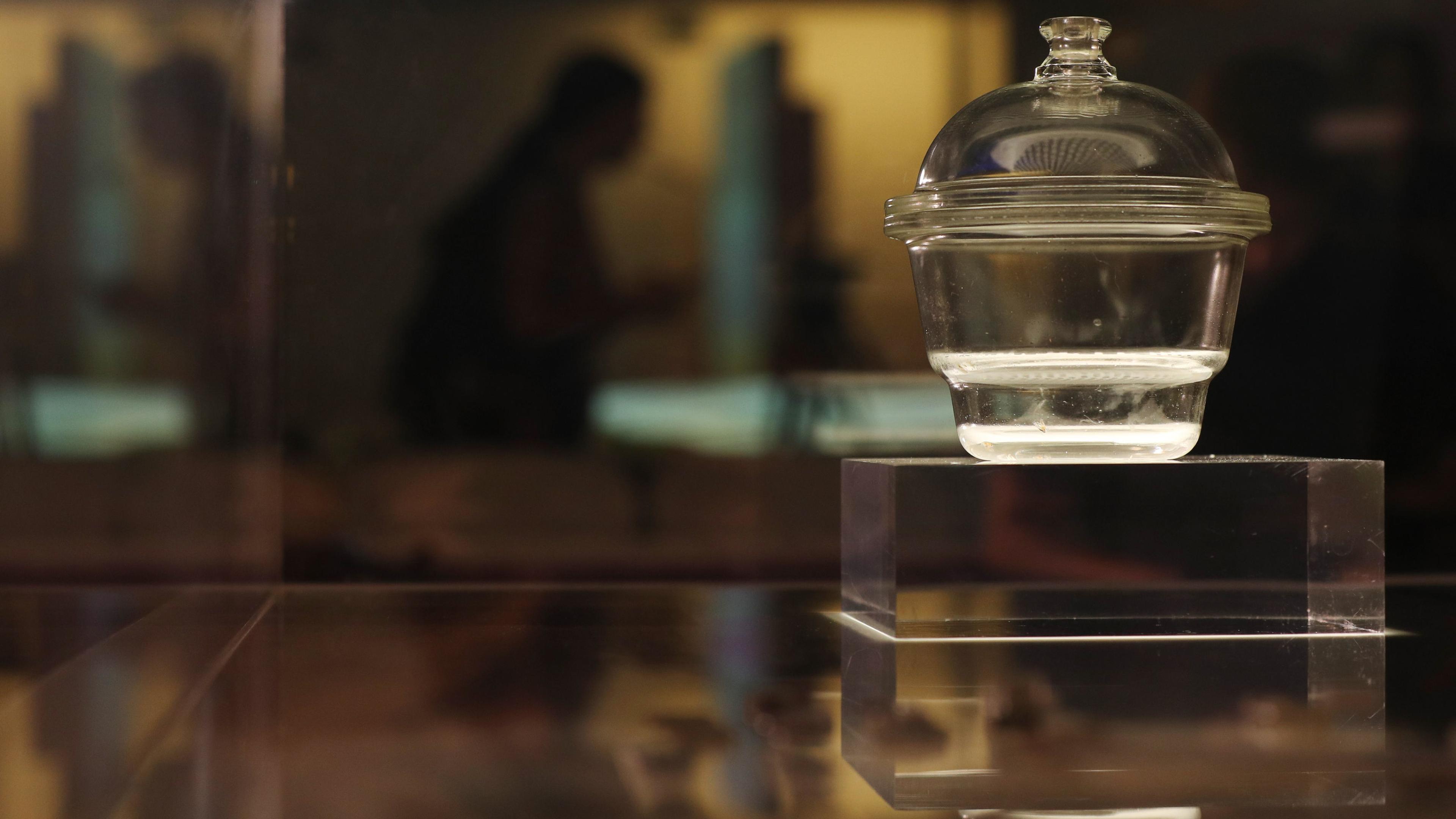 Photo of a glass jar with a lid on a pedestal in a dimly lit setting, with blurred figures in the background.