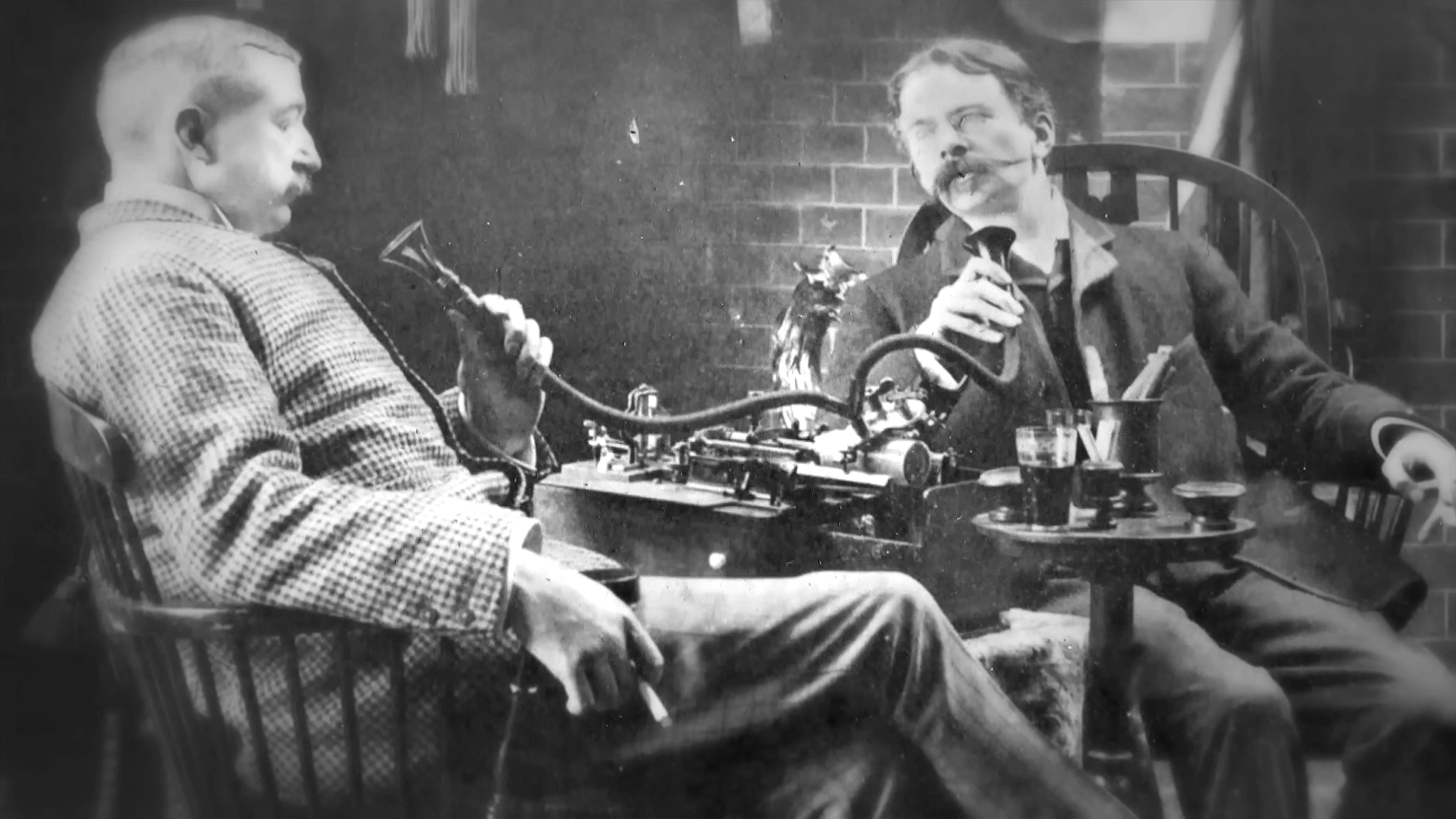 Black and white photo of two men using an early voice recording device, seated and holding speaking tubes.