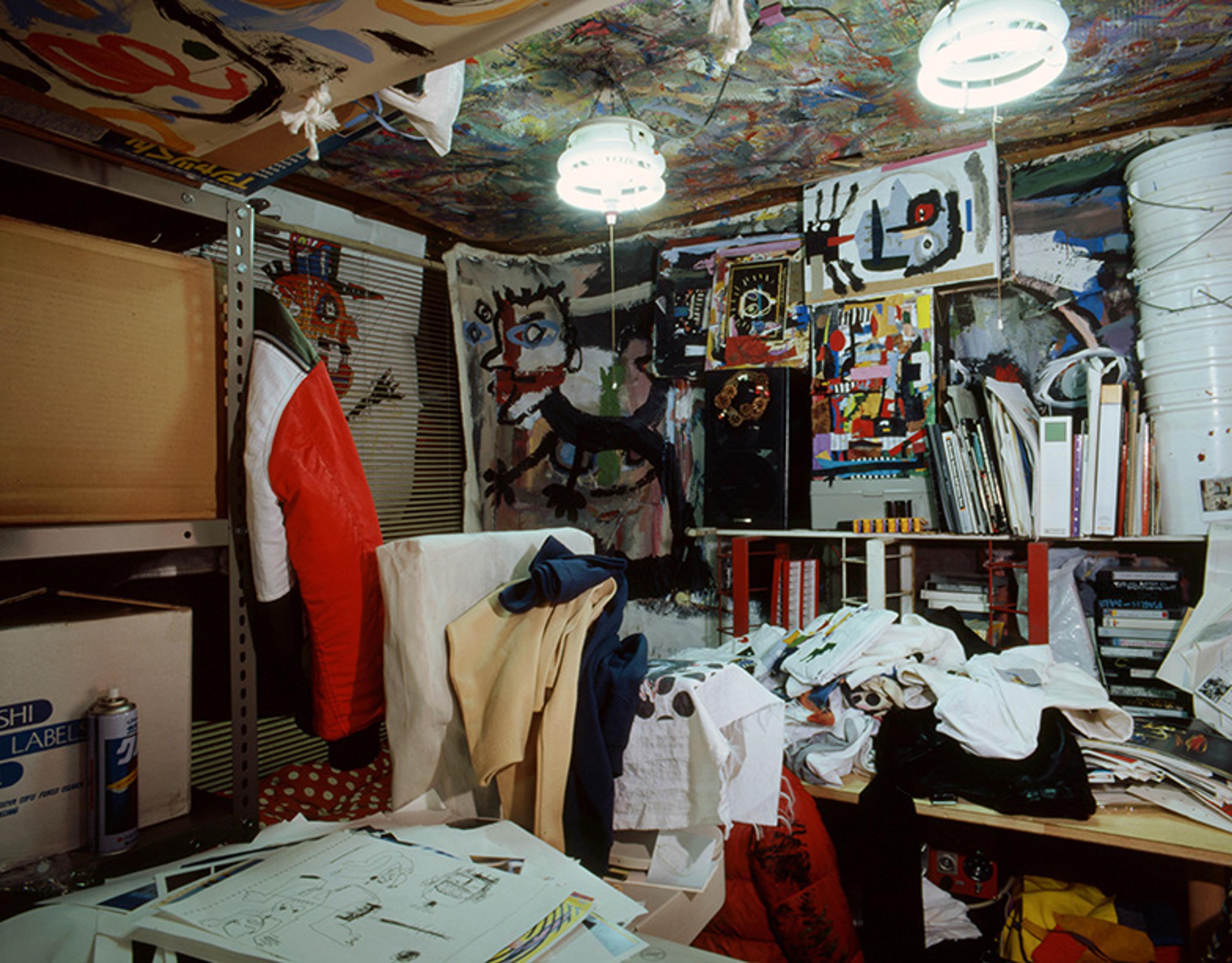 A cluttered art studio filled with paintings, clothes, books and papers, with a painted ceiling.