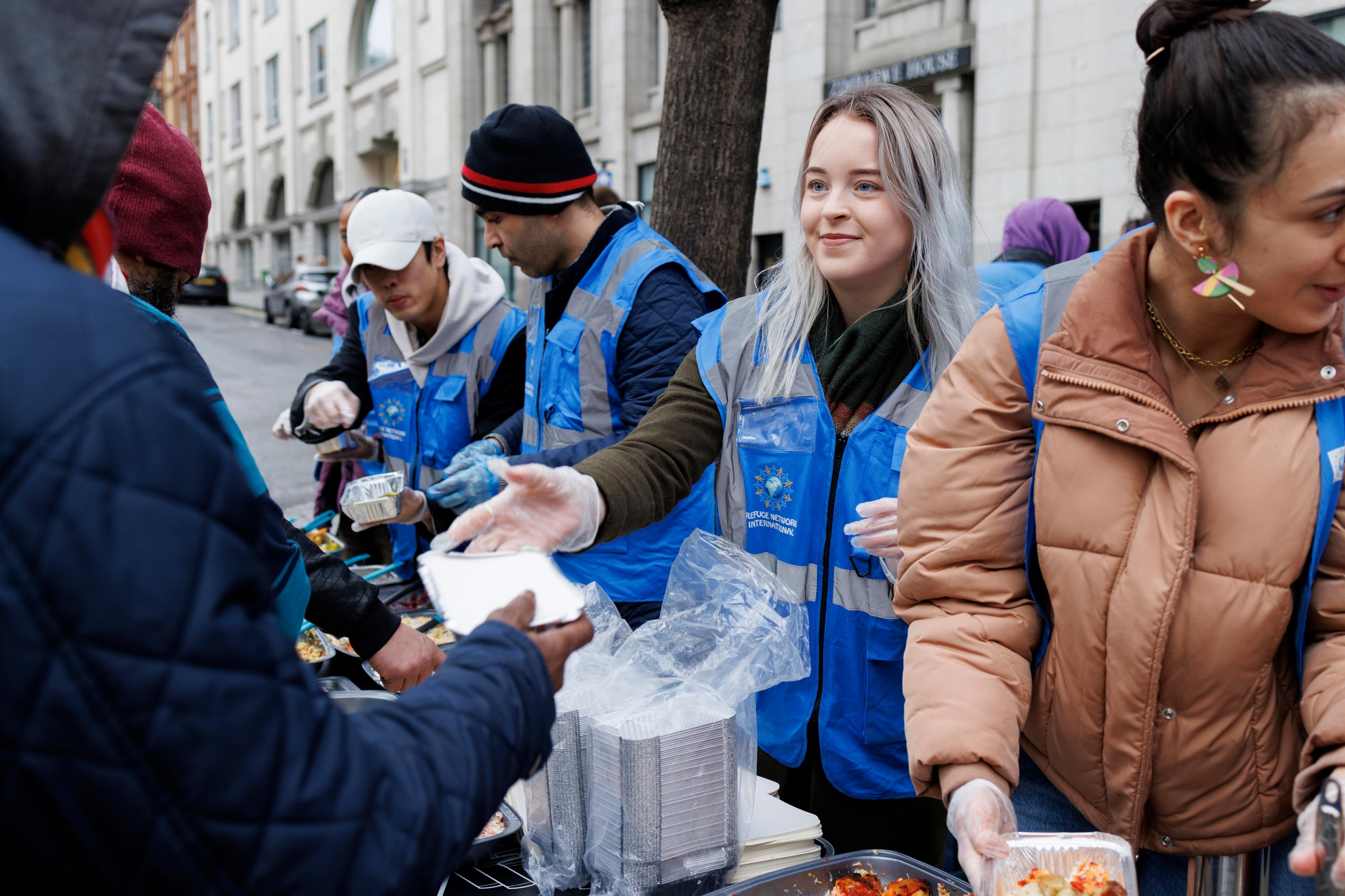 Photo of volunteers in blue vests distributing food to people on a city street, wearing gloves and standing by trays of food.