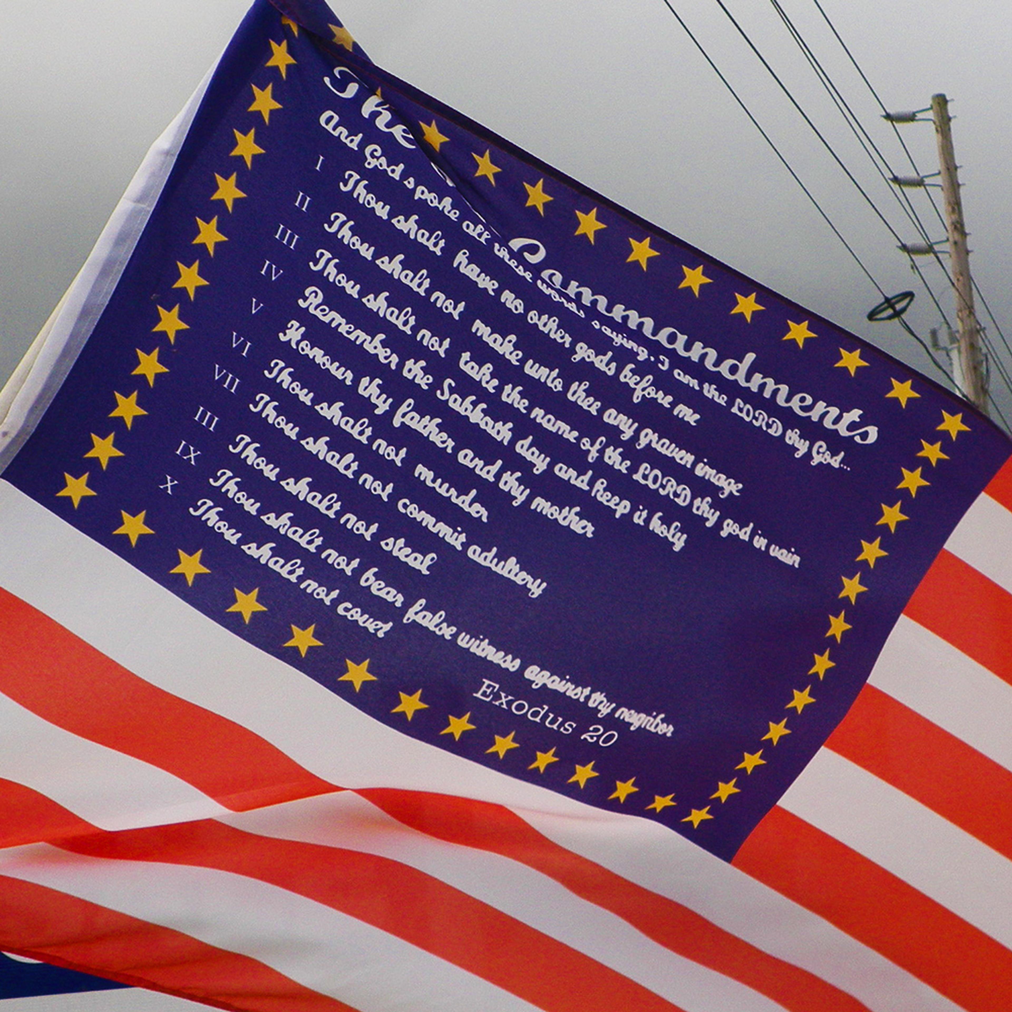 A US flag featuring the Ten Commandments in white text on a blue field with yellow stars, above red and white stripes.