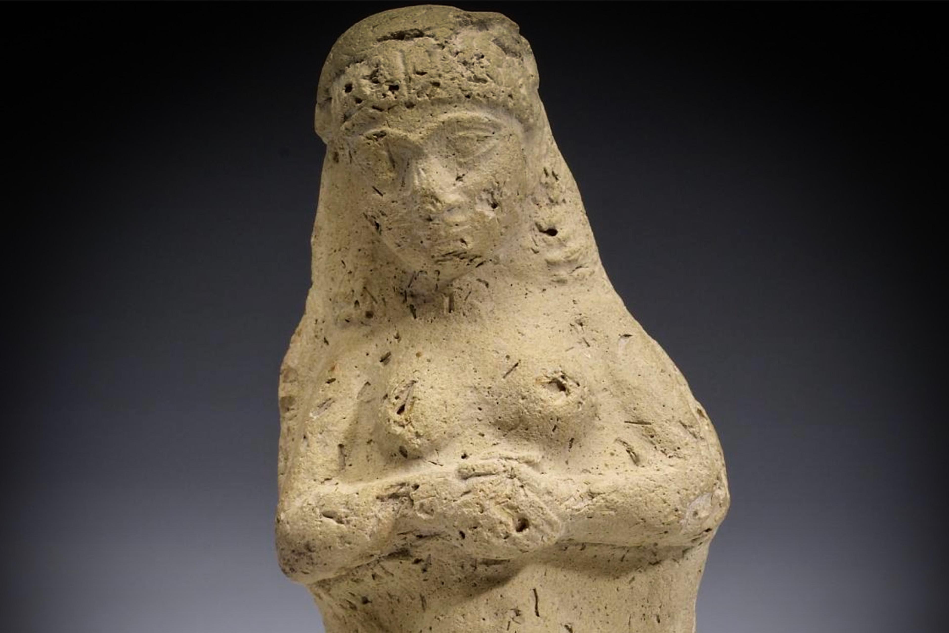 Ancient clay figurine of a woman with a veil, arms crossed. The surface is textured. Image shows a simple, worn design.