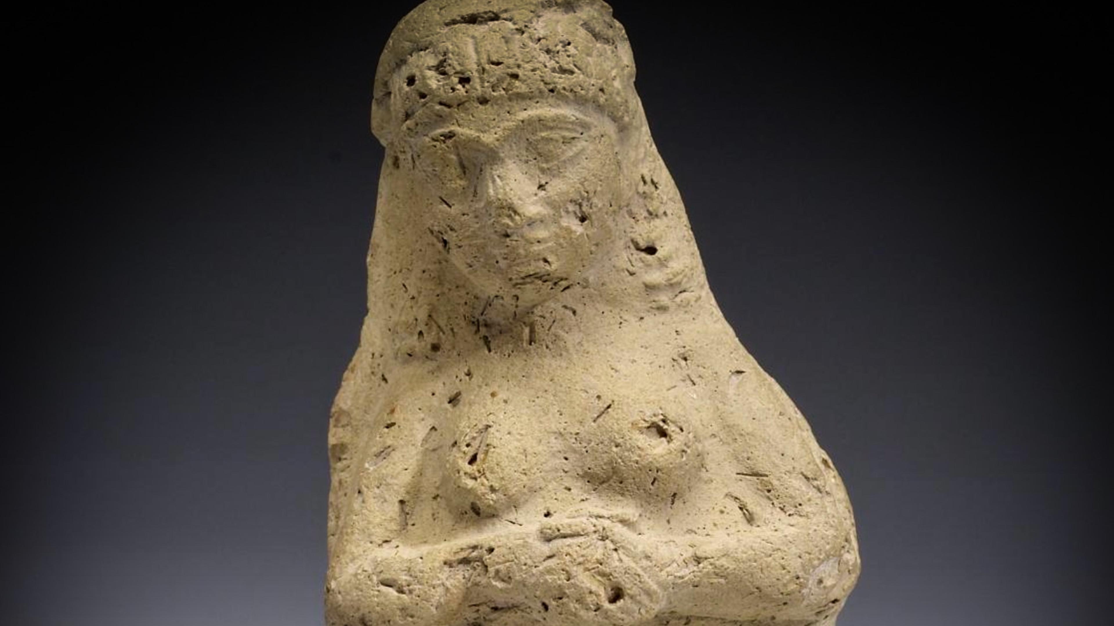 Ancient clay figurine of a woman with a veil, arms crossed. The surface is textured. Image shows a simple, worn design.