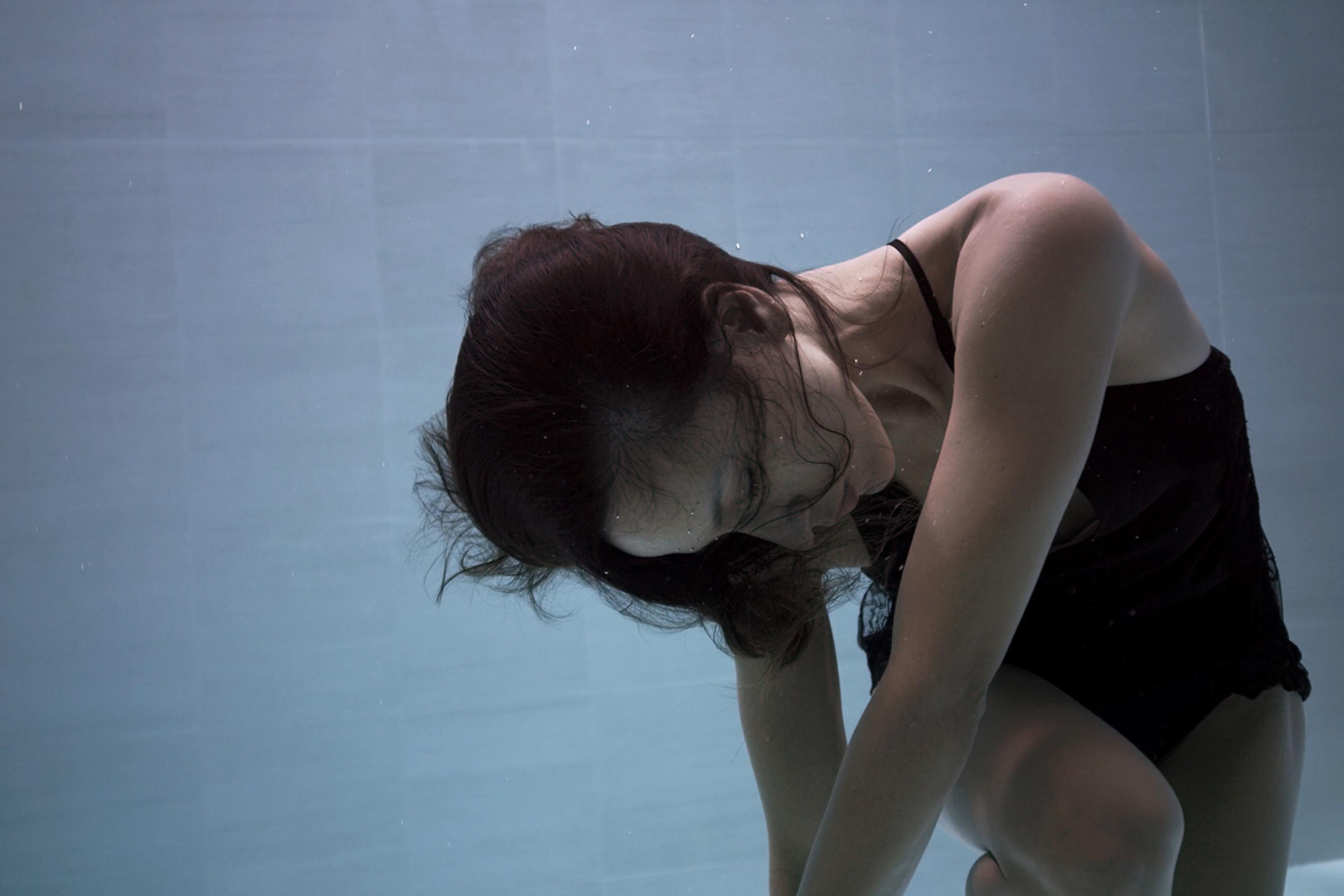 A woman in a black swimsuit is submerged underwater, with her head down and hair floating gently around her.