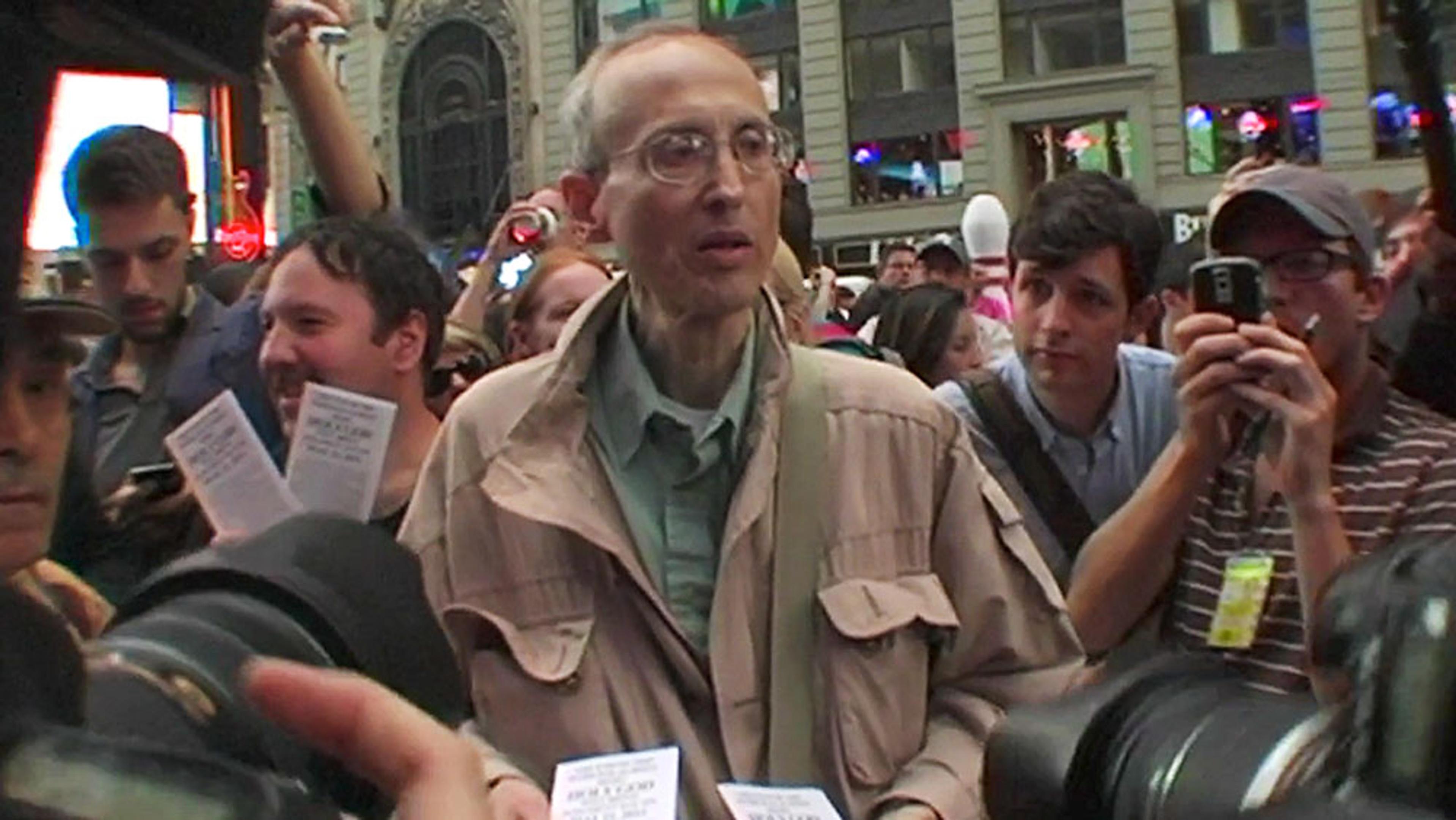 A man in a taupe jacket holding papers, surrounded by a crowd with cameras and phones, set against an urban background.