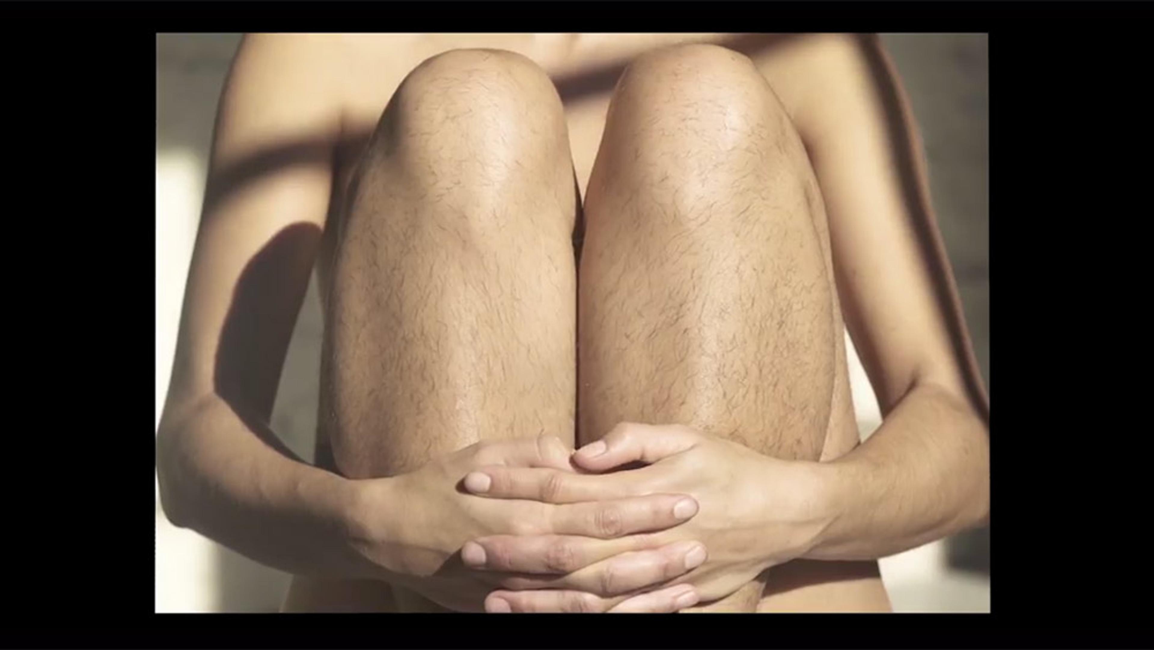 A woman with hairy legs sitting with her knees up, arms wrapped around her calves, in soft lighting. Subtle shadows visible.