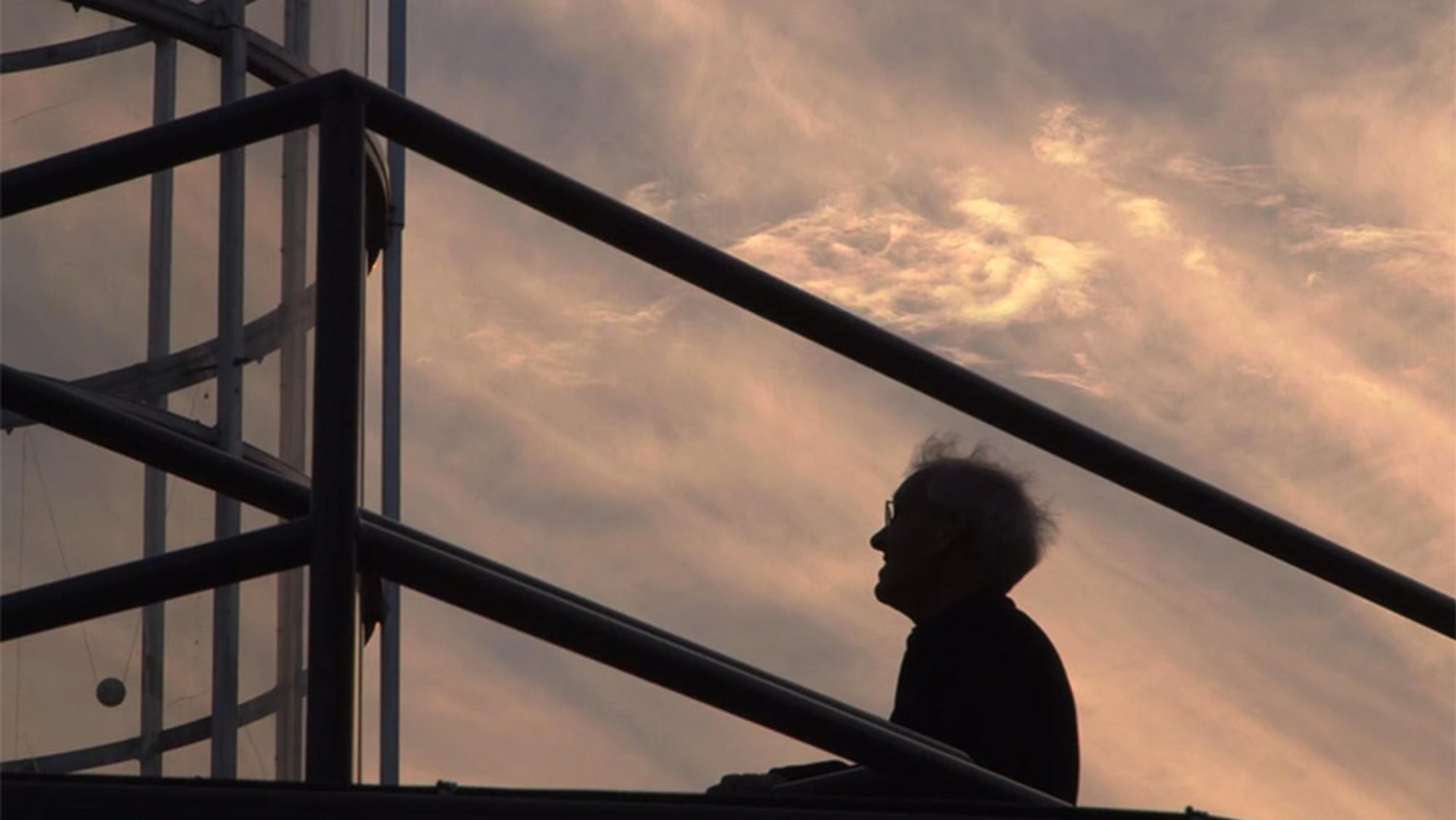 A man in silhouette on a staircase, with railings and a cloudy sky backdrop. The scene appears during sunset or sunrise.
