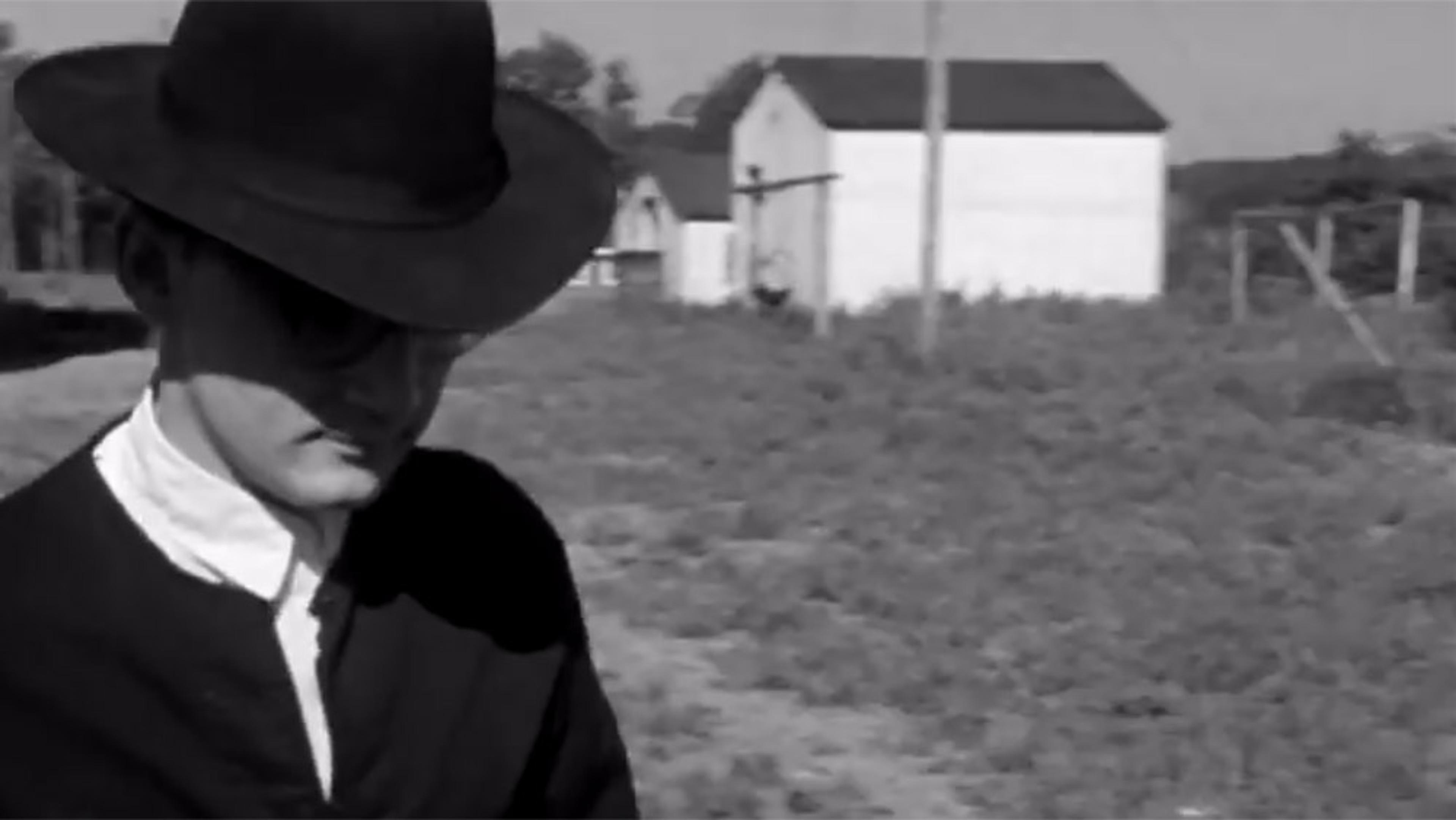 Black-and-white photograph of a person wearing a dark hat and coat in a rural area, with blurred buildings and a field in the background.