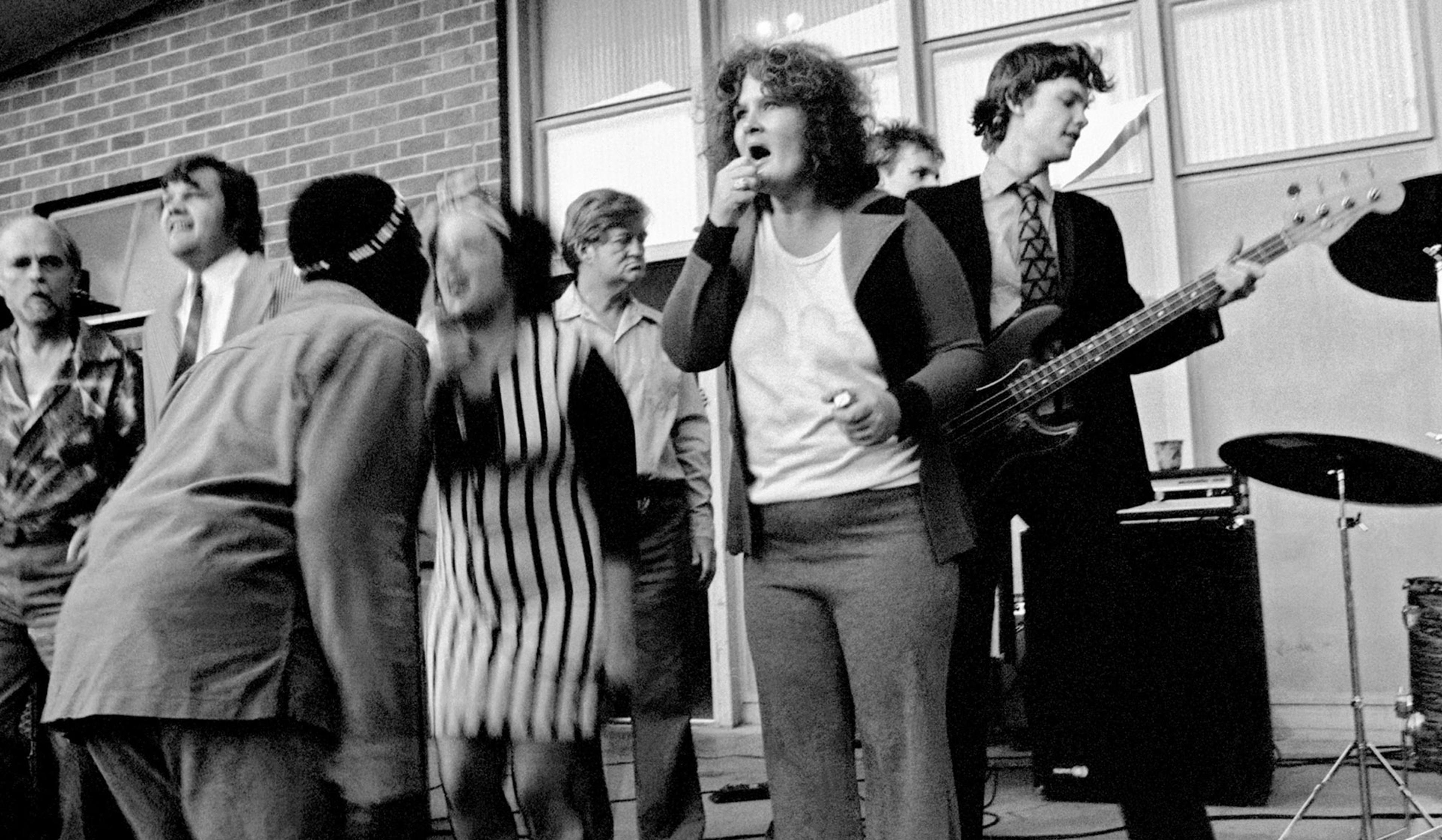 Black-and-white photo of a band performing on stage surrounded by a confused chaos of people.