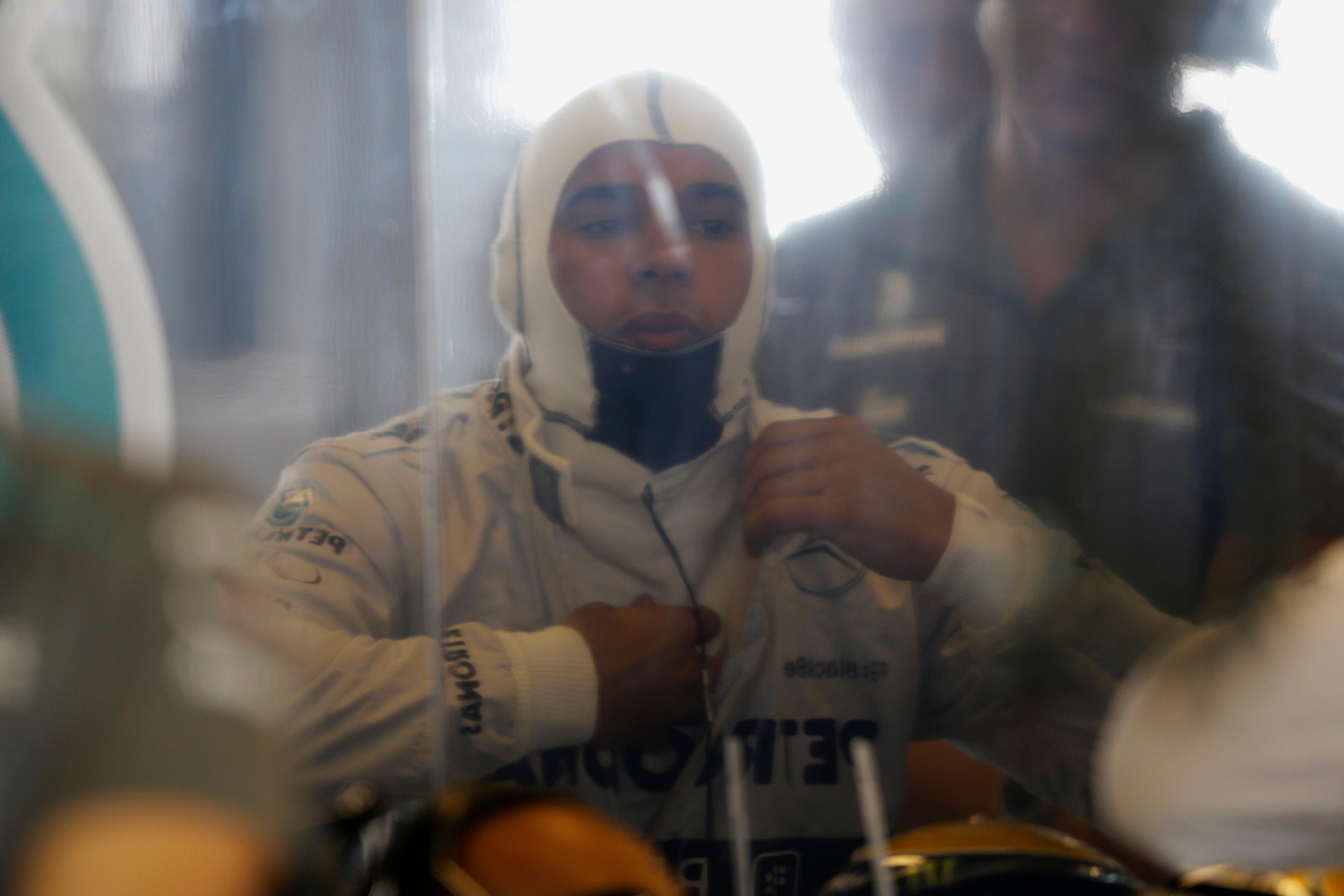 Photo of a racing driver in a helmet and suit, adjusting gear, seen through a reflection or a glass surface.