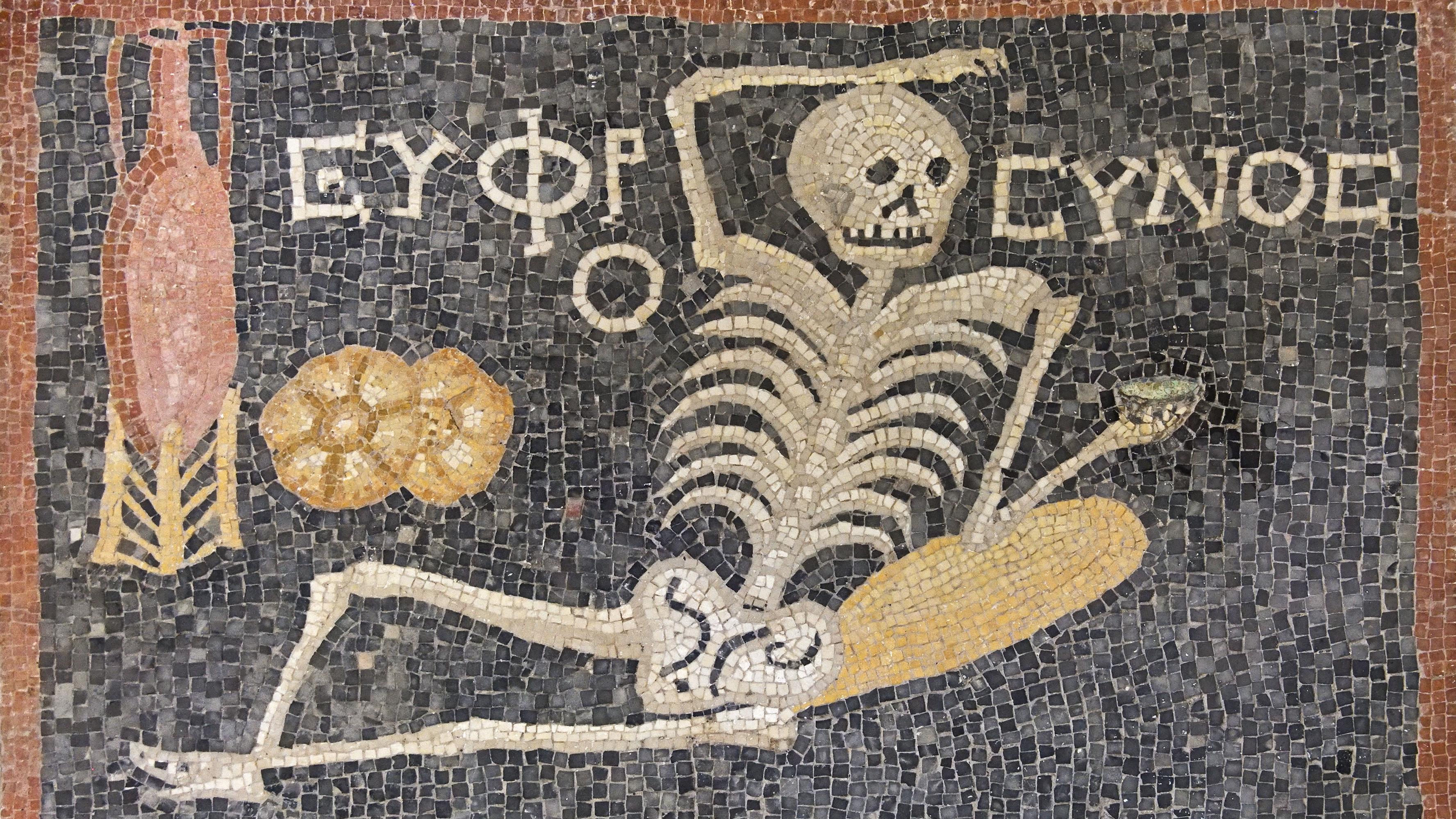 Ancient mosaic of a reclining skeleton with Greek text, holding a drink, next to an amphora and bread on a dark background.