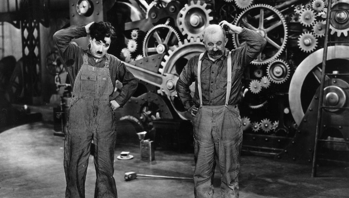 Charlie Chaplin and Chester Conklin, dressed in overalls, scratch their heads, standing in front of large industrial gears and machinery in a black-and-white scene.