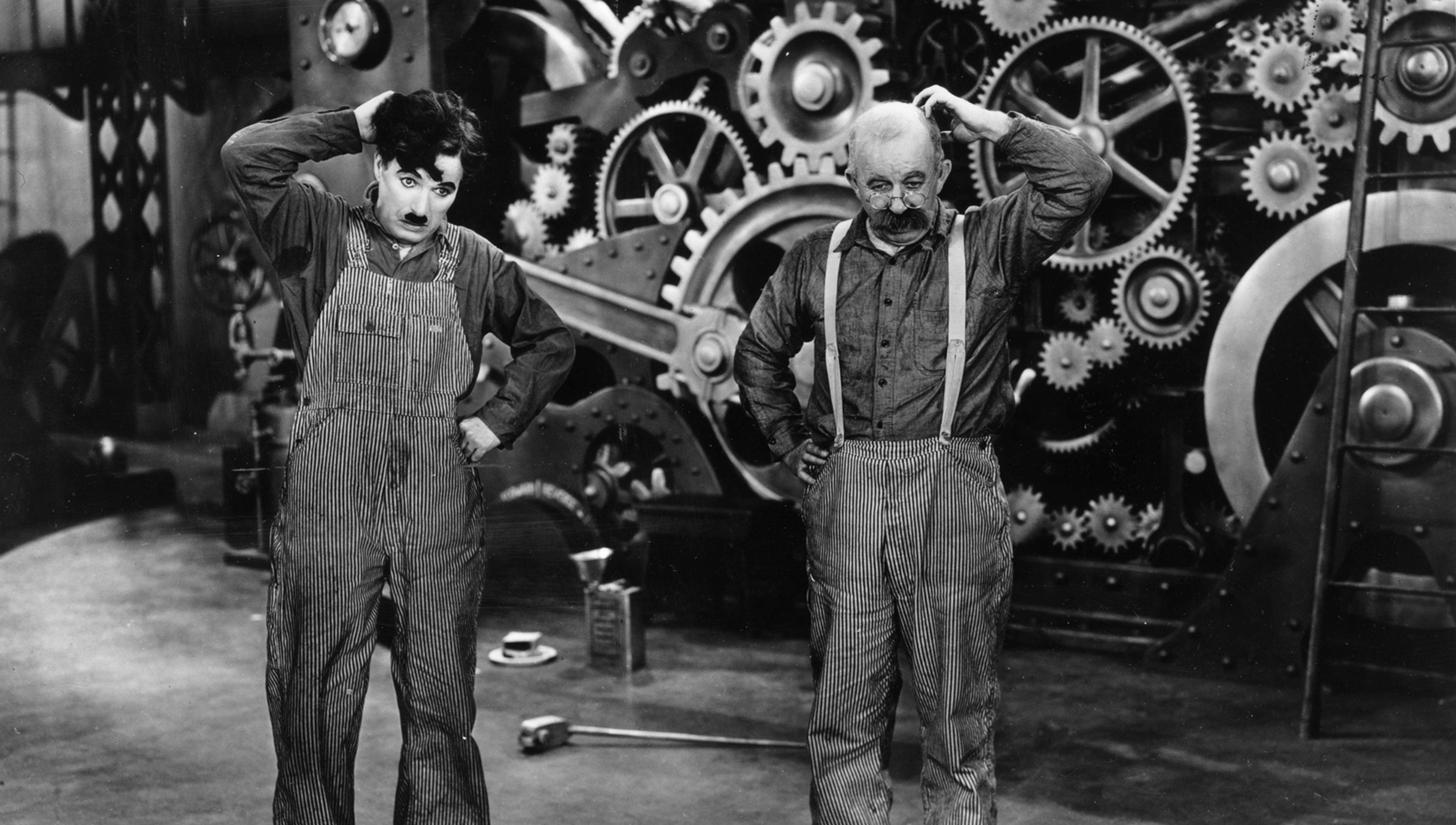 Charlie Chaplin and Chester Conklin, dressed in overalls, scratch their heads, standing in front of large industrial gears and machinery in a black-and-white scene.