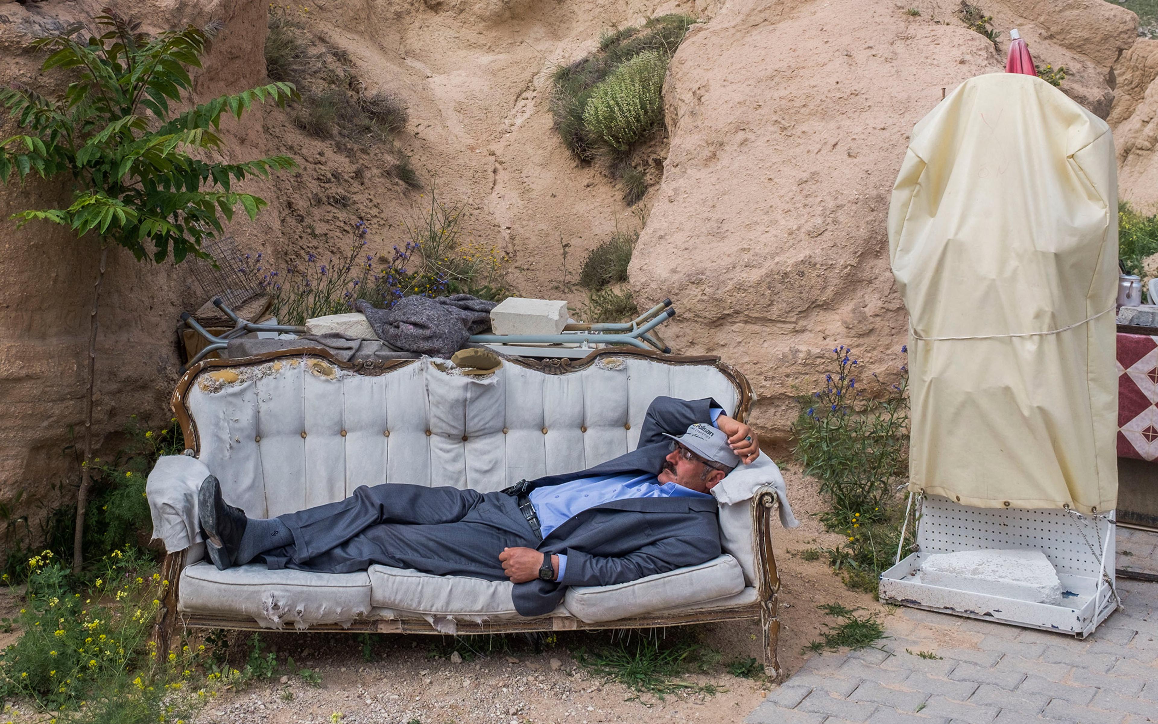 A man in a suit and cap lying on a tattered sofa outdoors next to a rocky hillside with various items scattered around.