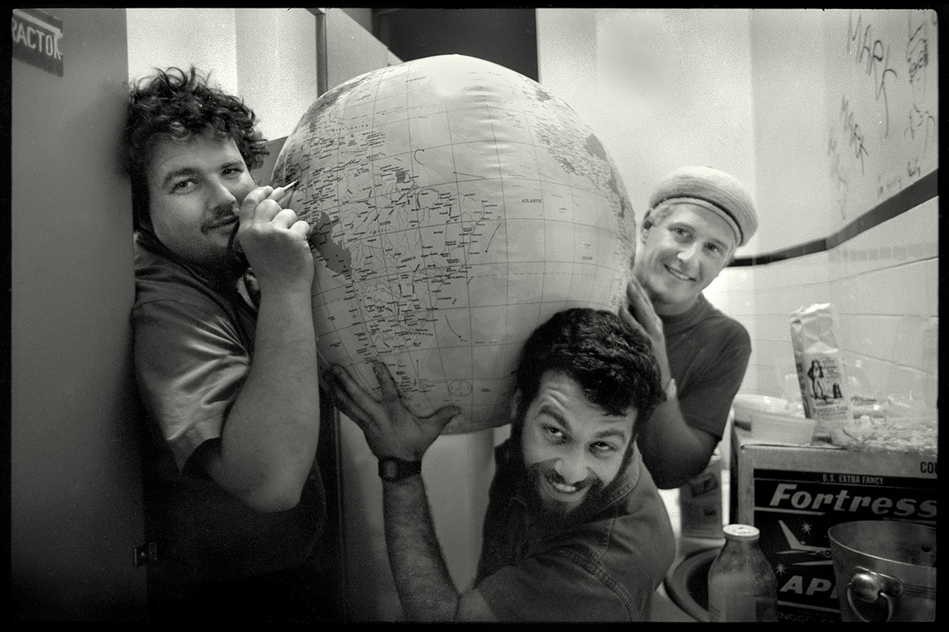 Photo of three men smiling and holding a large inflatable globe in a room with various boxes and jars.