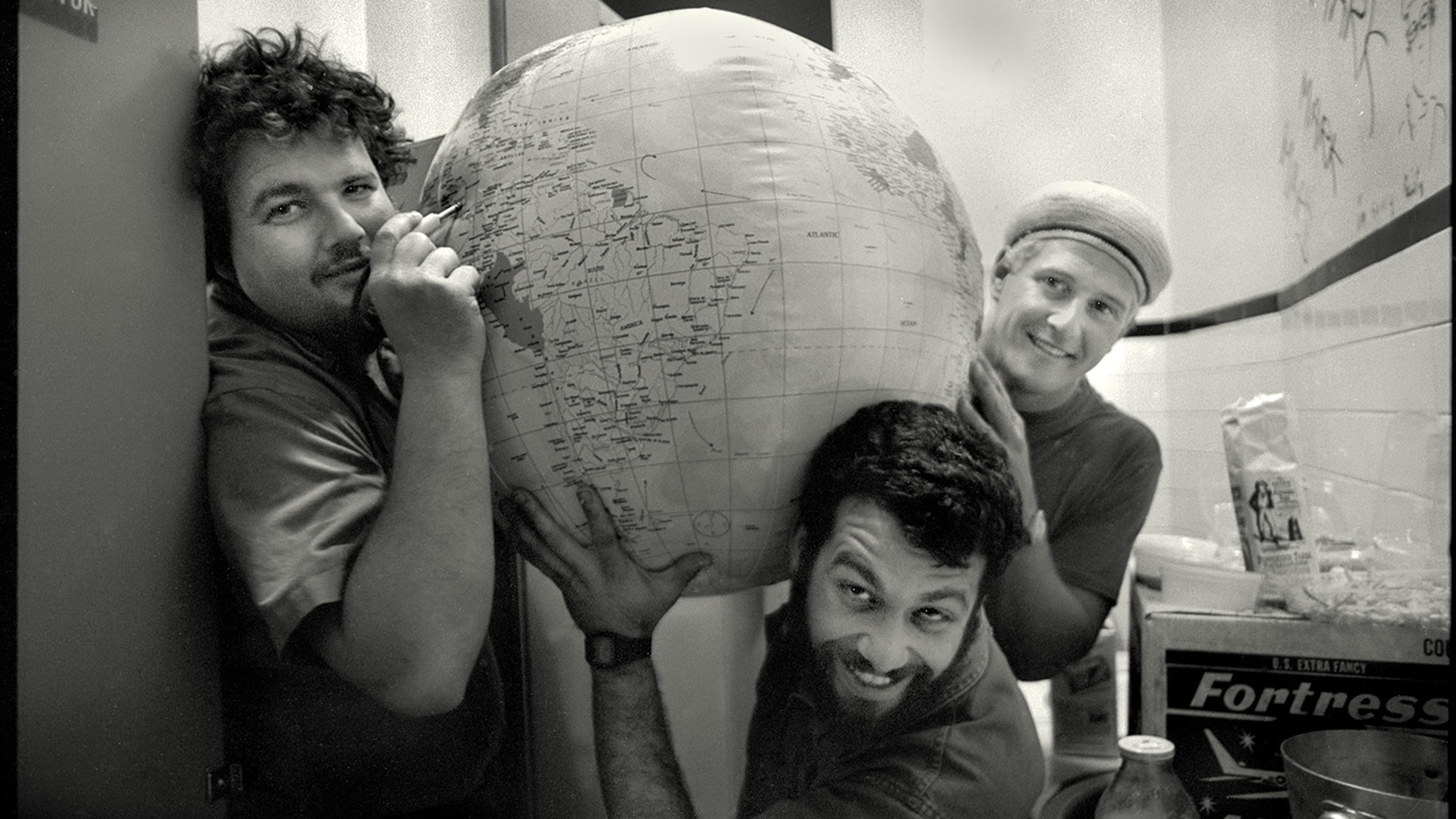 Photo of three men smiling and holding a large inflatable globe in a room with various boxes and jars.