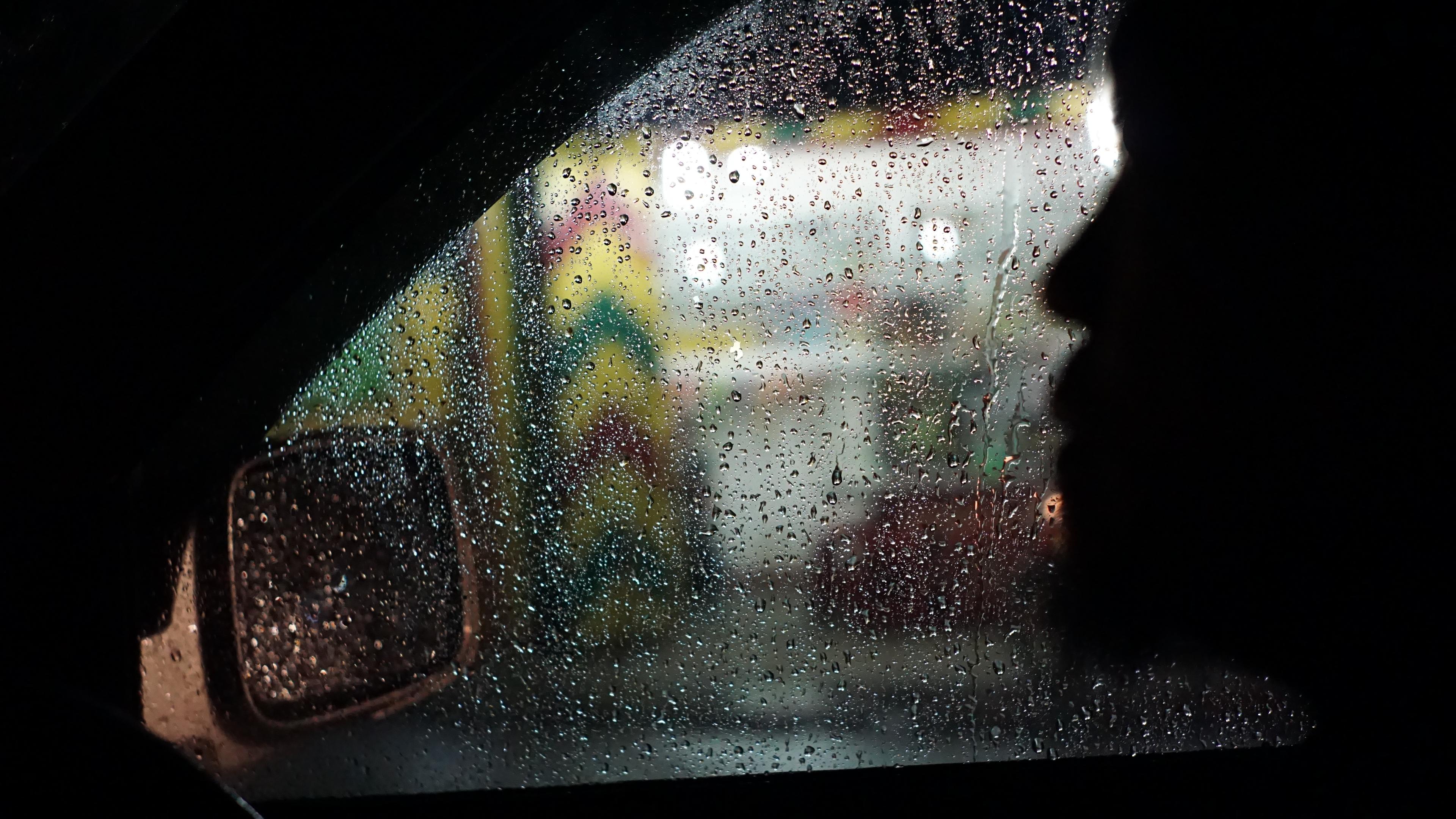 Photo of a rainy car window with a blurred face silhouette inside and colourful lights outside.