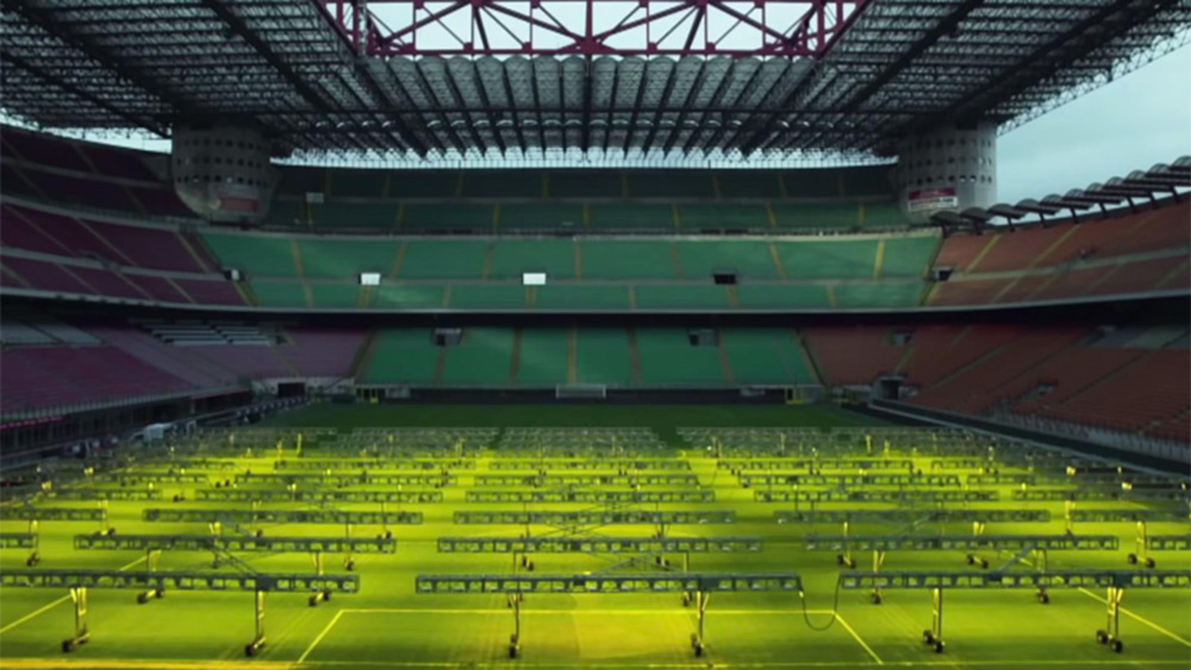 An empty football stadium with grow lights on the pitch, green and red seating areas, and an intricate roof structure