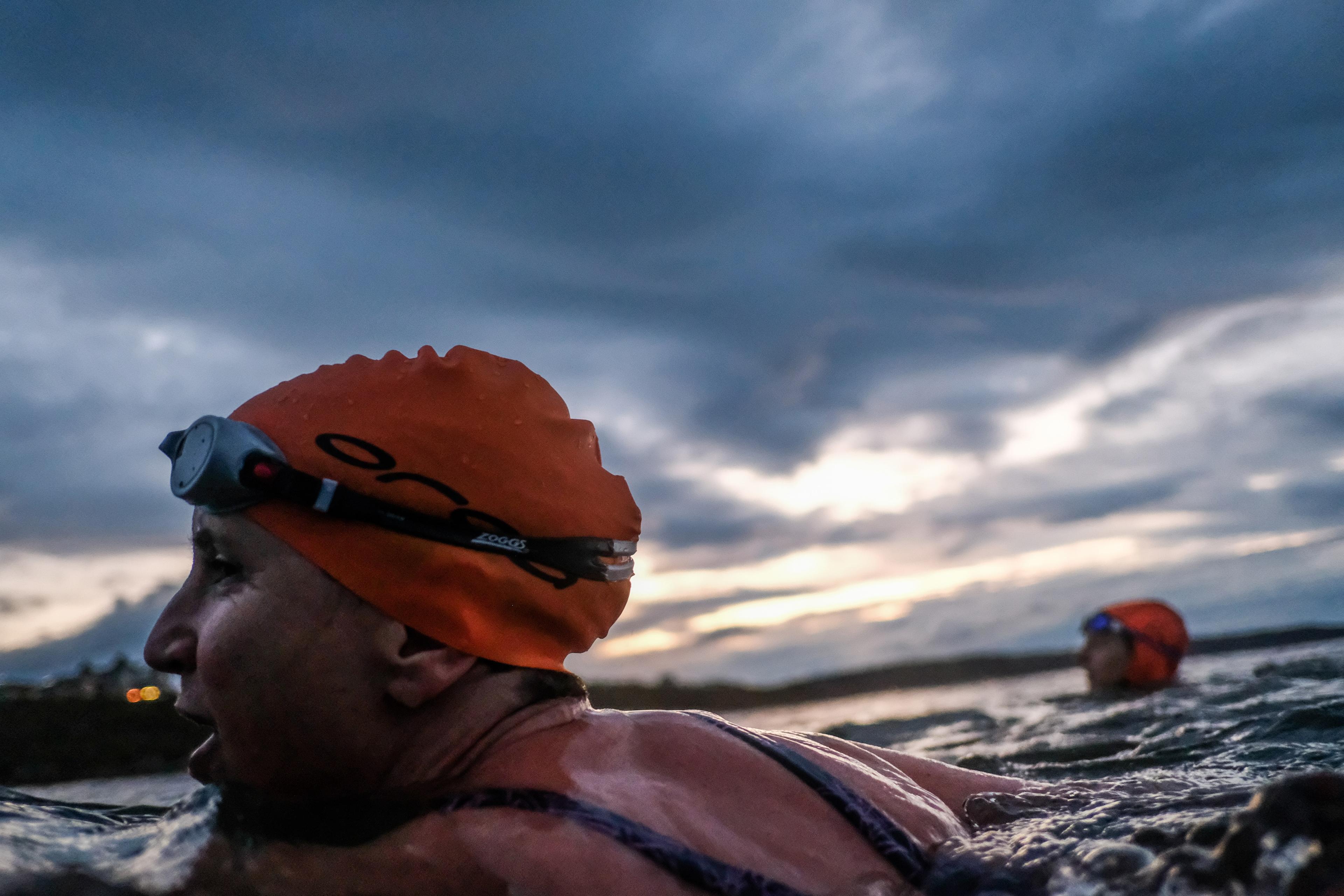 Photo of two swimmers in orange caps at sunset in open water, with a cloudy sky in the background.
