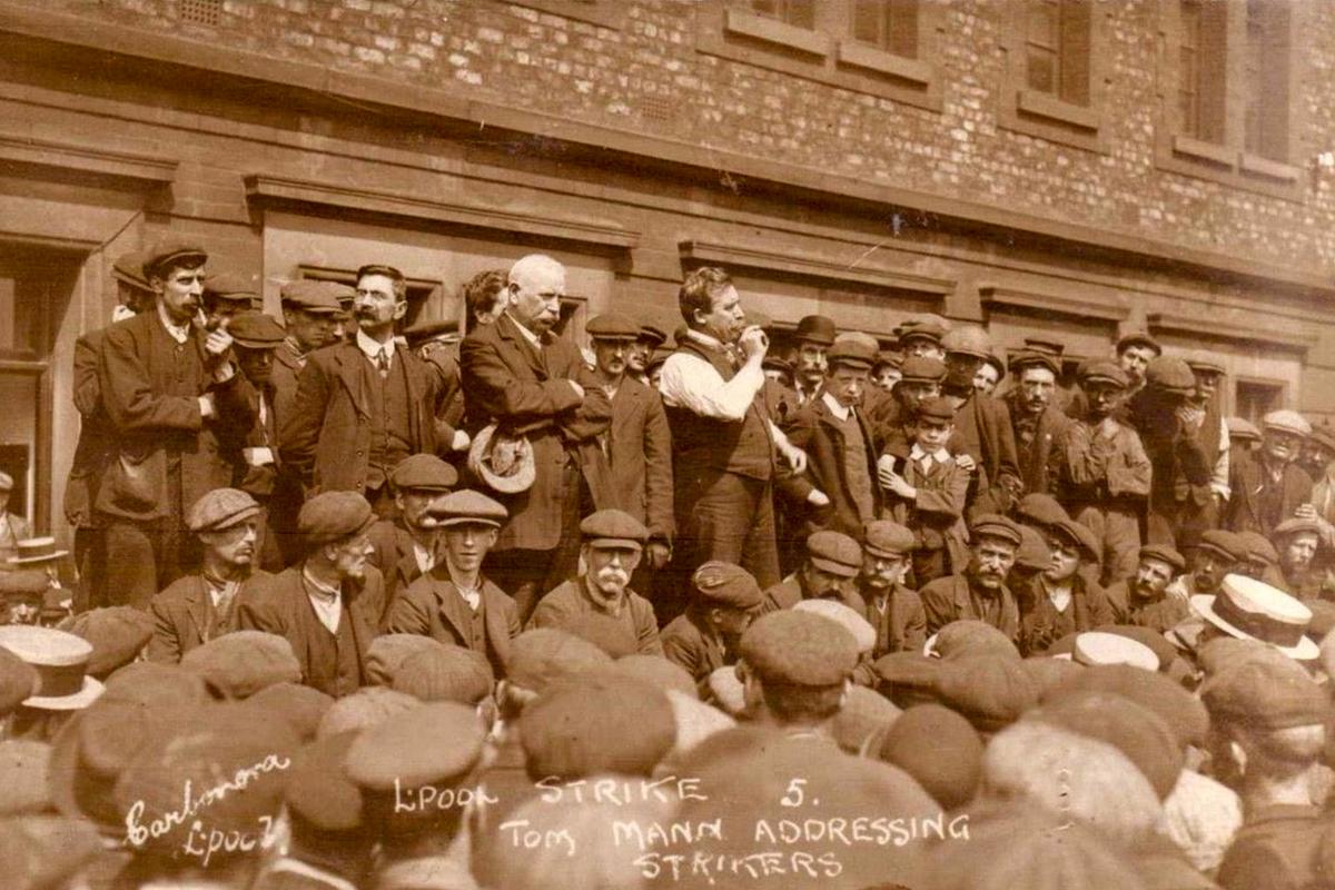 Sepia-toned photo of a man addressing a crowd of strikers outside a brick building wearing early 20th-century attire.