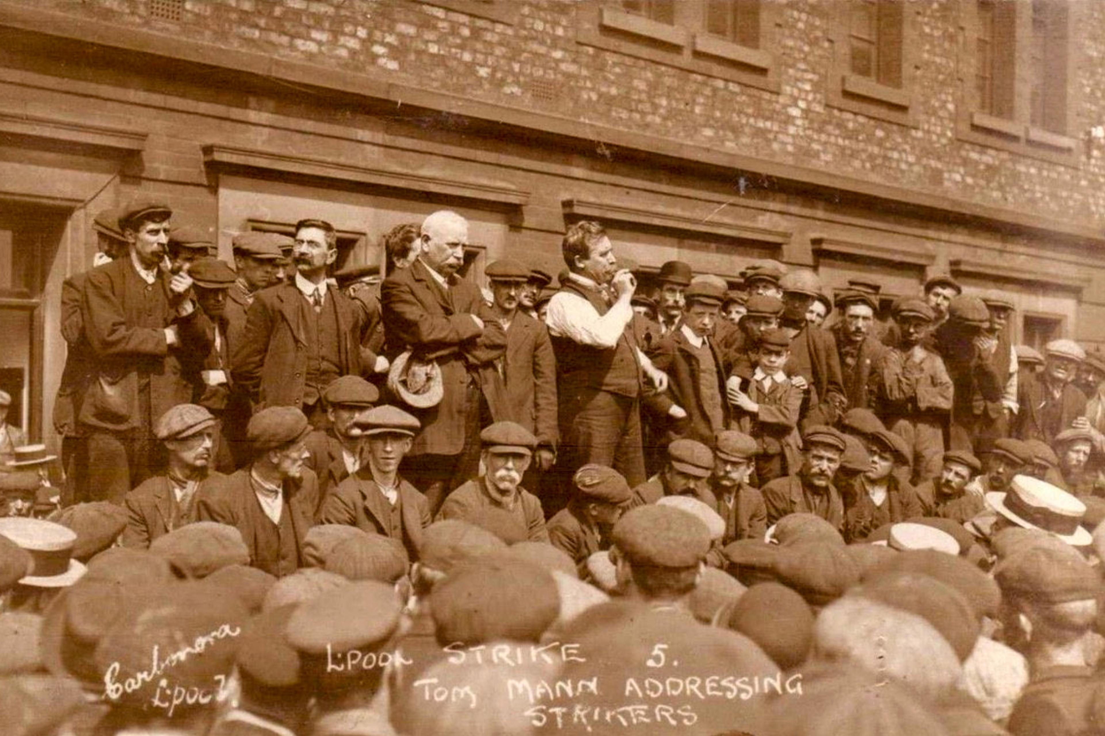 Sepia-toned photo of a man addressing a crowd of strikers outside a brick building wearing early 20th-century attire.
