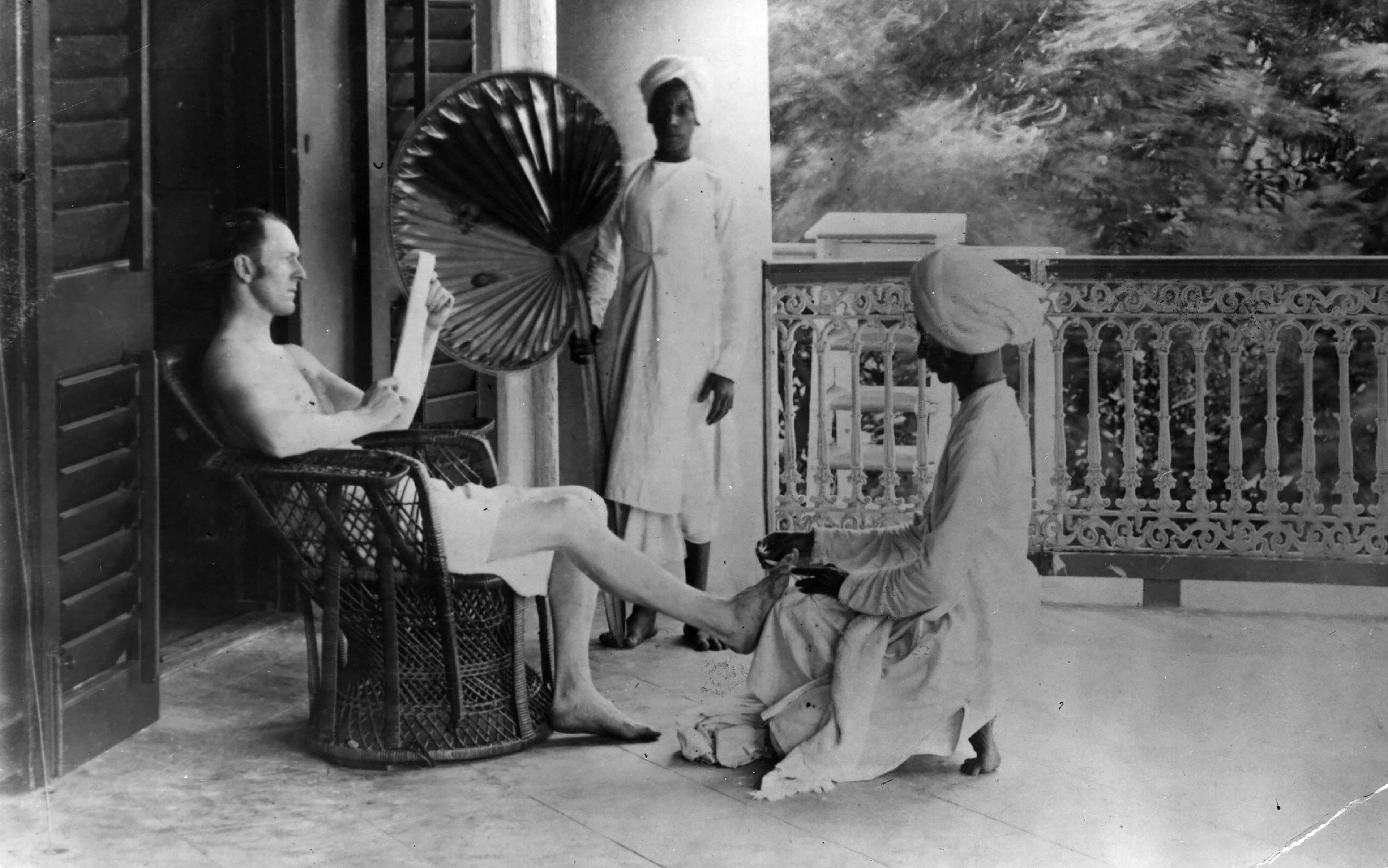 Black-and-white photo of a white man in a wicker chair on a veranda attended by two Black servants, one holding a fan and the other massaging his feet.