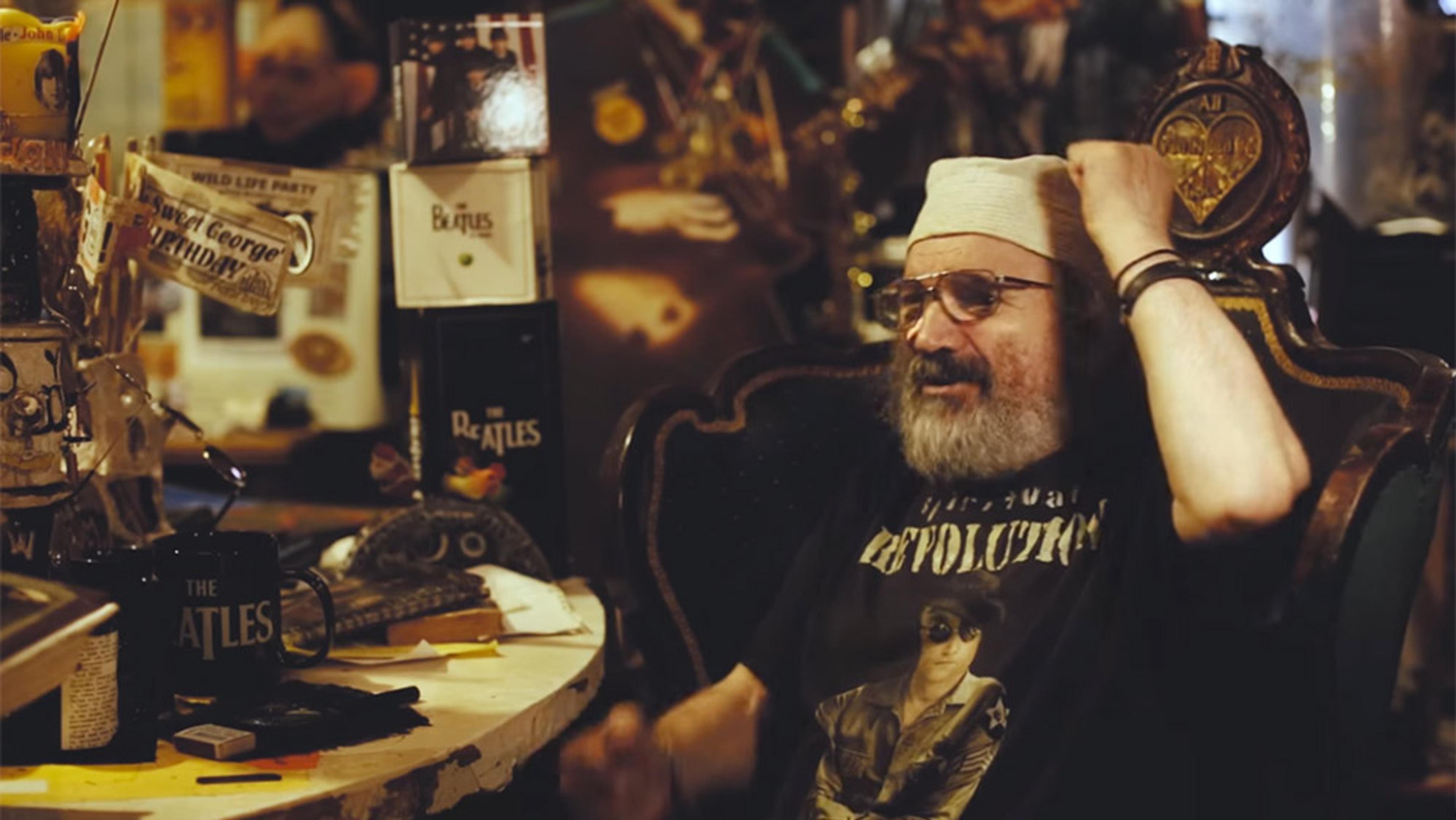 A bearded older man in glasses and a hat sits in a cluttered room filled with Beatles memorabilia, including mugs and posters.