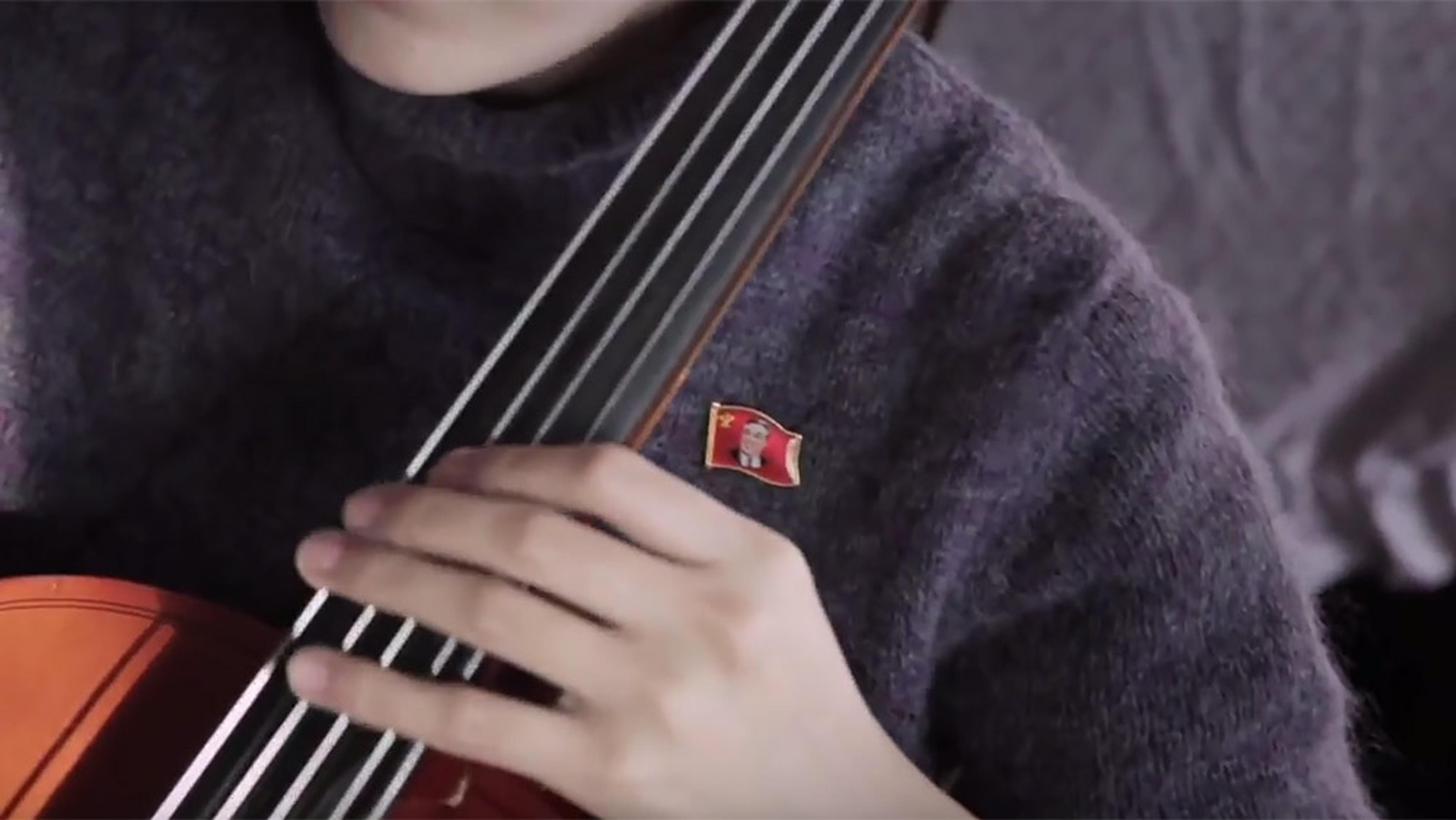 A person playing a cello, their hand on the fingerboard, wearing a grey sweater with a red badge pinned to it.