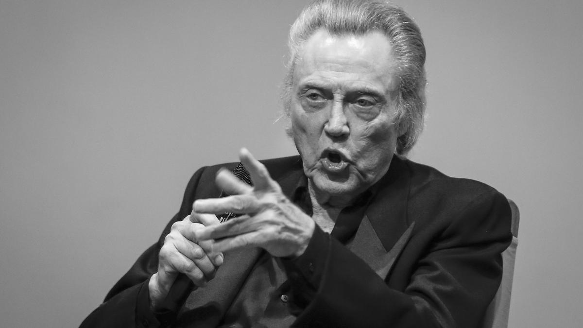 Black and white photo of an older man who is Christopher Walken in a suit gesturing while speaking into a microphone.