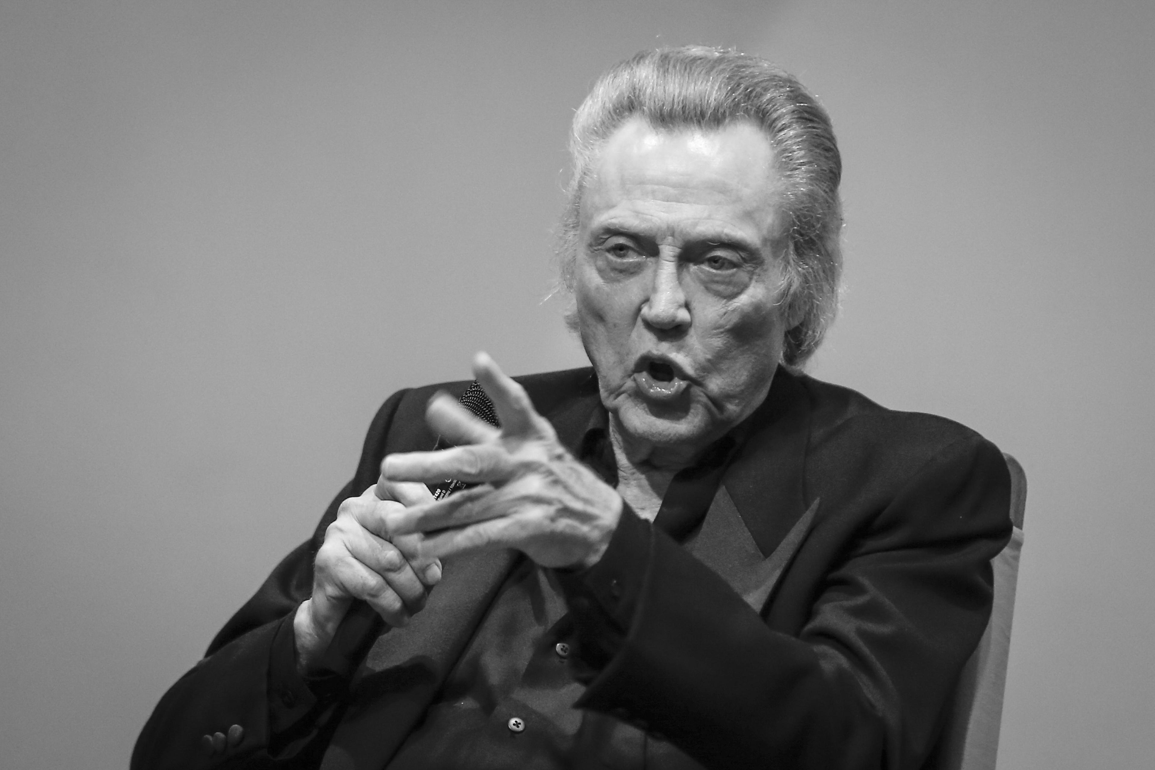 Black and white photo of an older man who is Christopher Walken in a suit gesturing while speaking into a microphone.