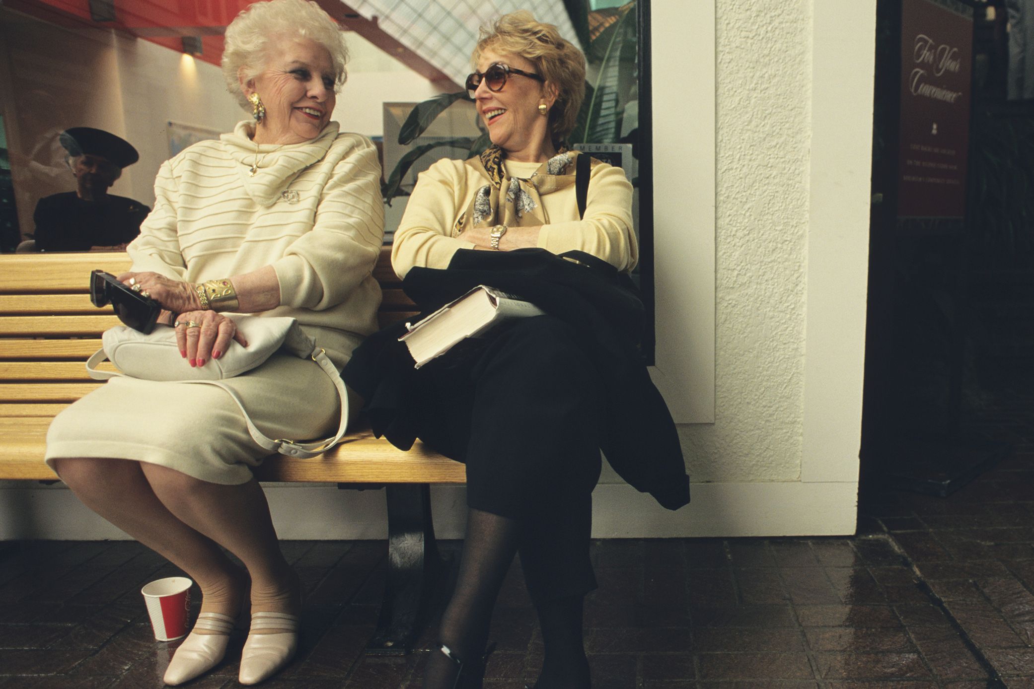 Two older women sitting on a bench, smiling at each other, one holding a book.