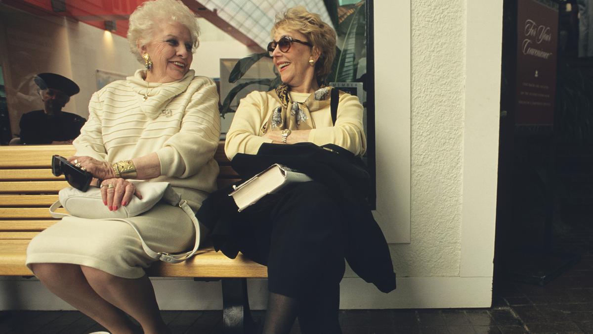 Two older women sitting on a bench, smiling at each other, one holding a book.