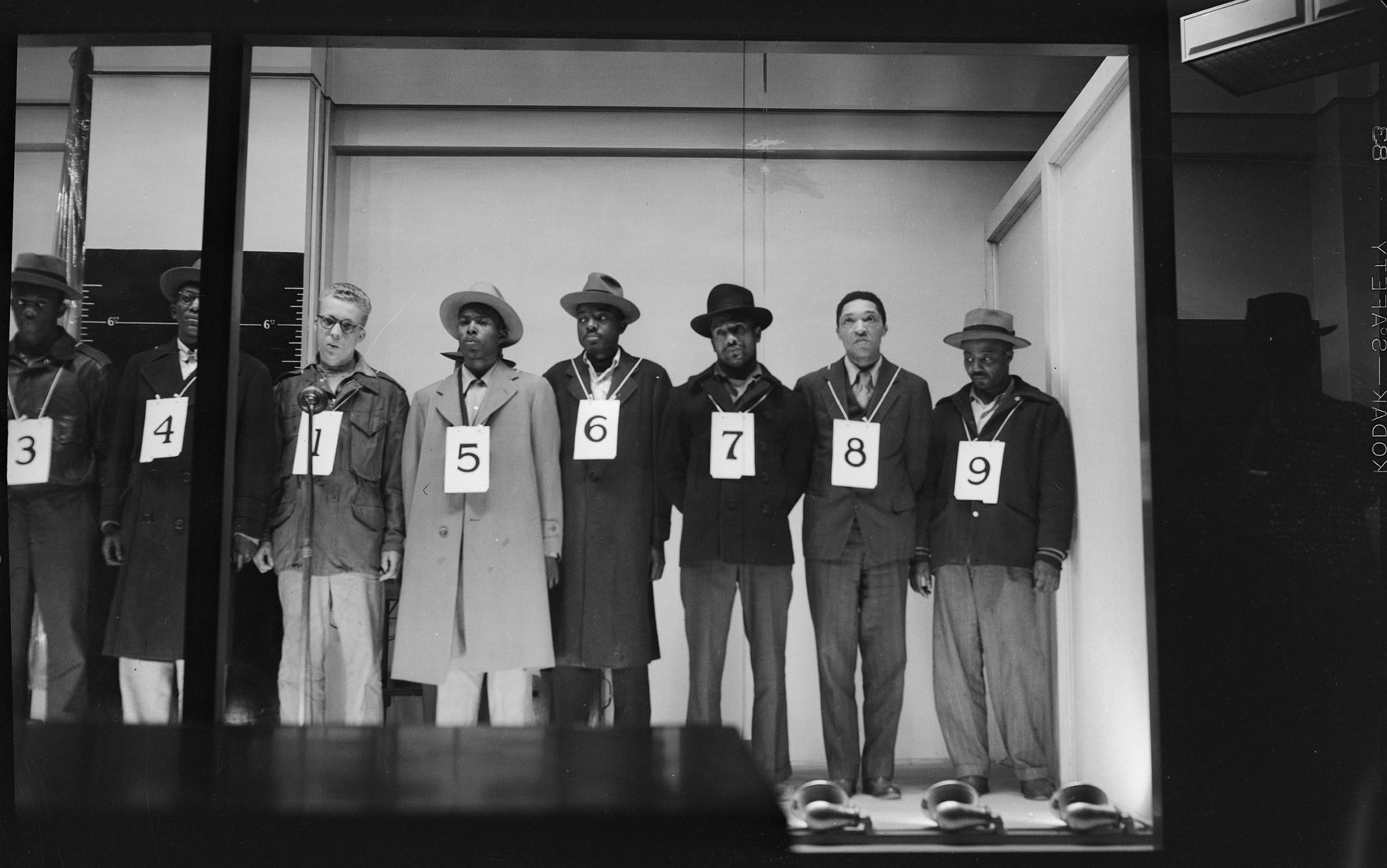 Black and white photo of seven Black men and one white man in an identification line-up wearing numbered signs and facing forward.