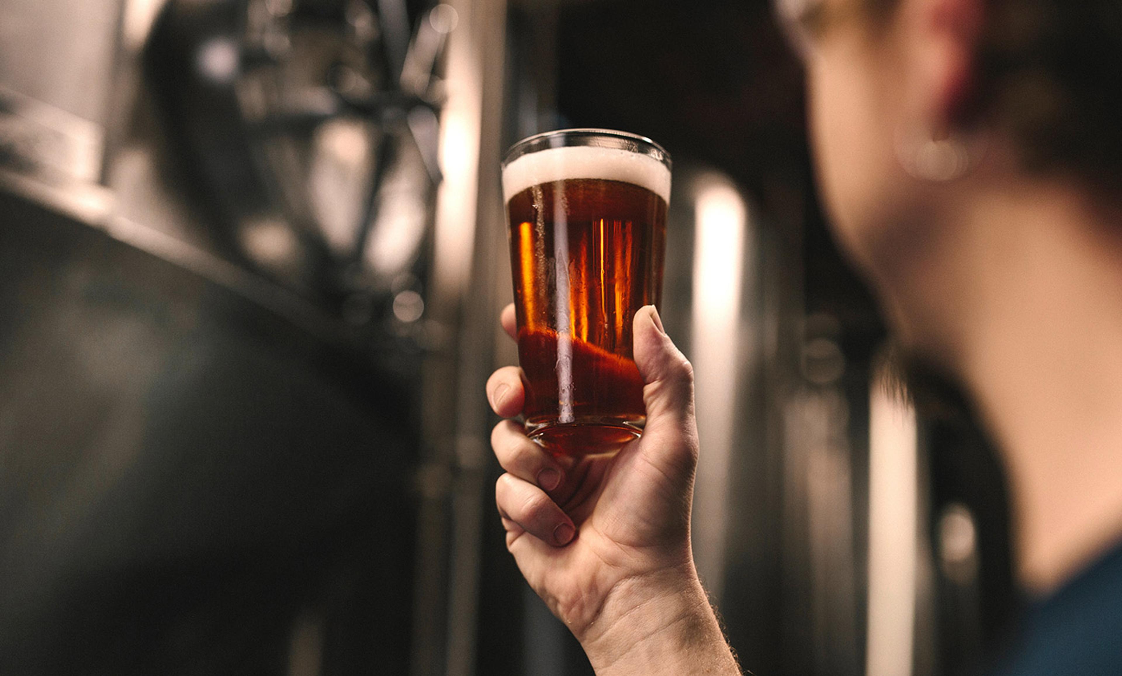 A person holding up a glass of amber-coloured beer in a brewery, with brewing equipment visible in the background.