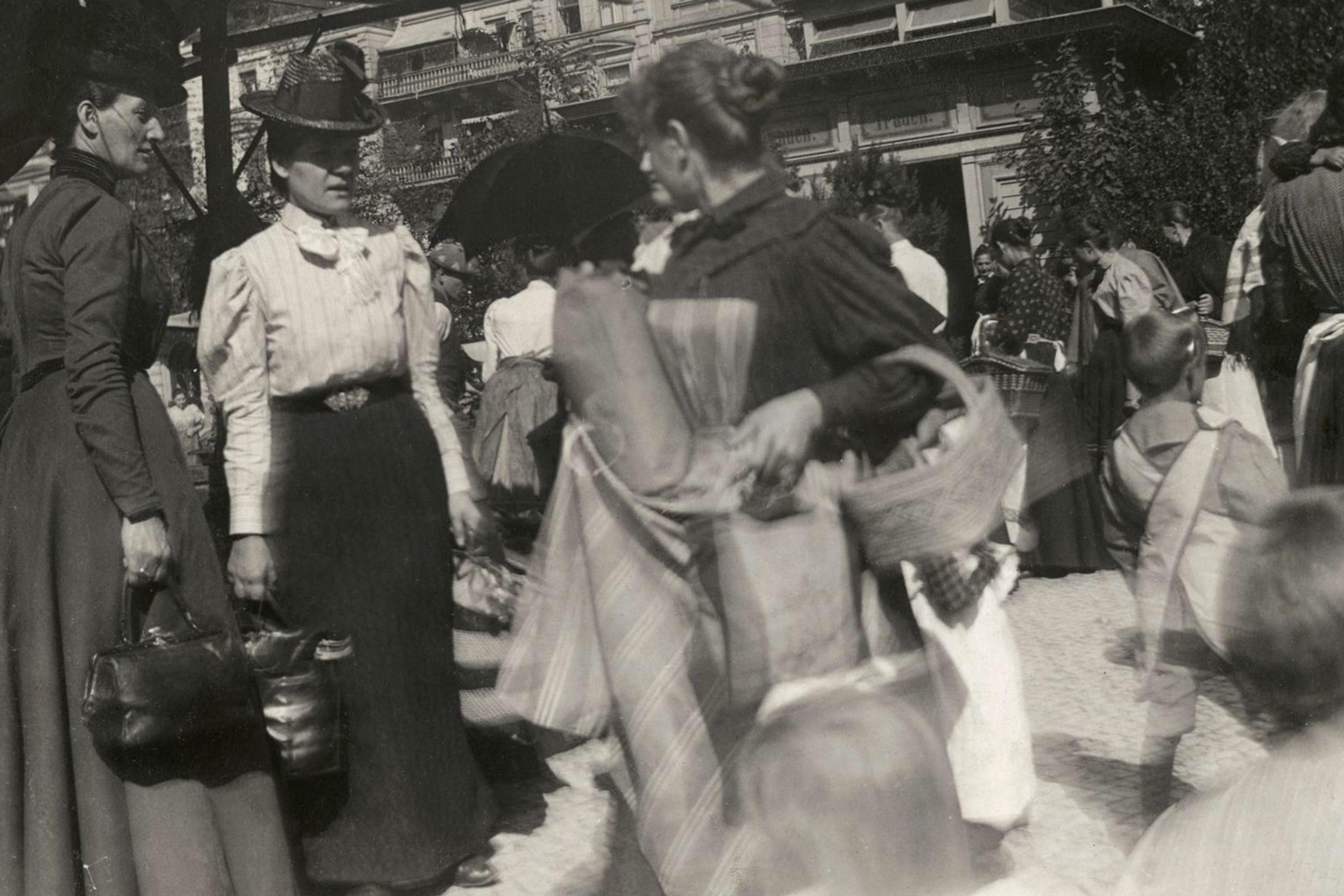 A vintage photo showing people dressed in old-fashioned clothing, carrying baskets and bags, walking through a busy outdoor market area.