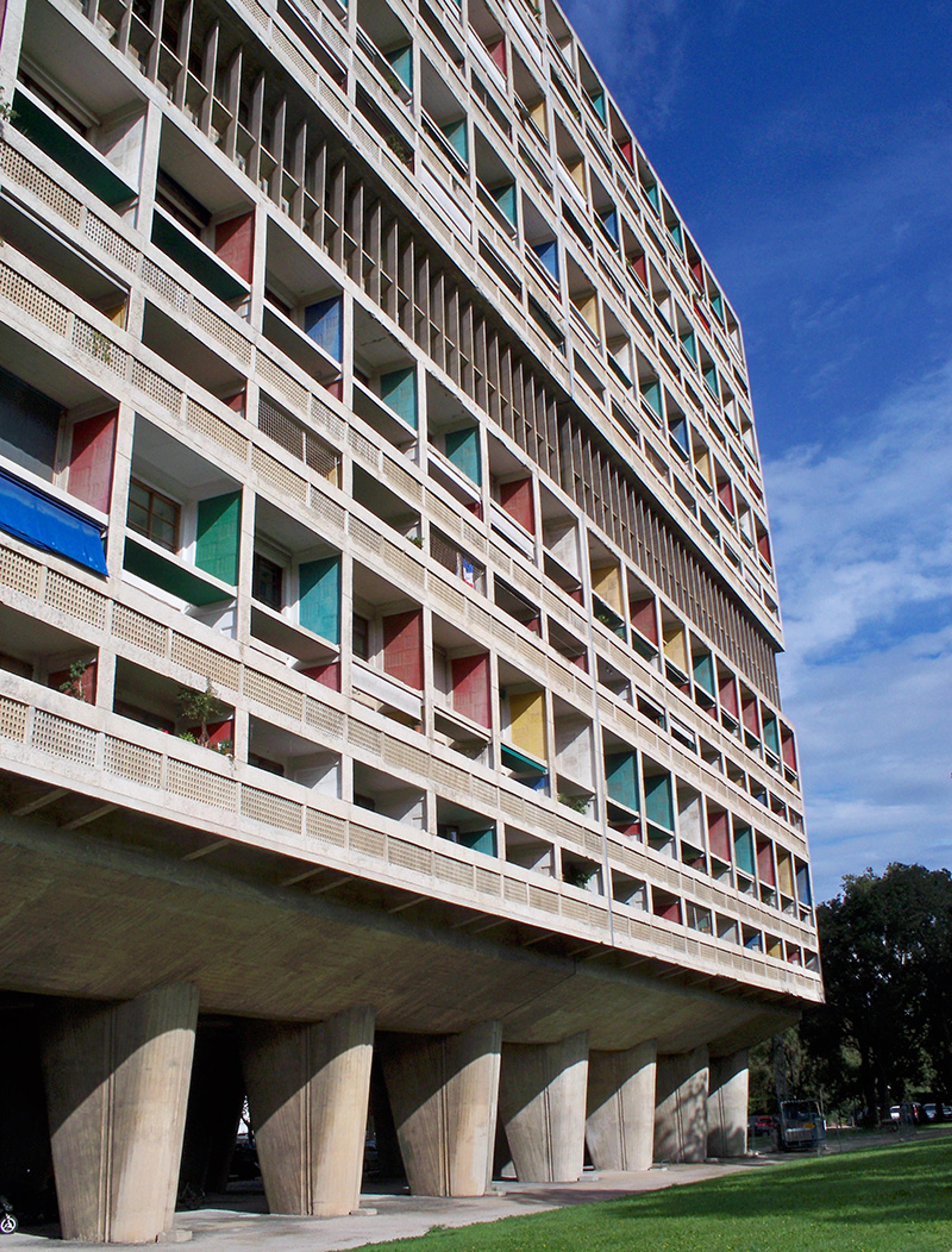 A modernist building with colourful balconies on large concrete pillars against a blue sky, viewed from a low angle.