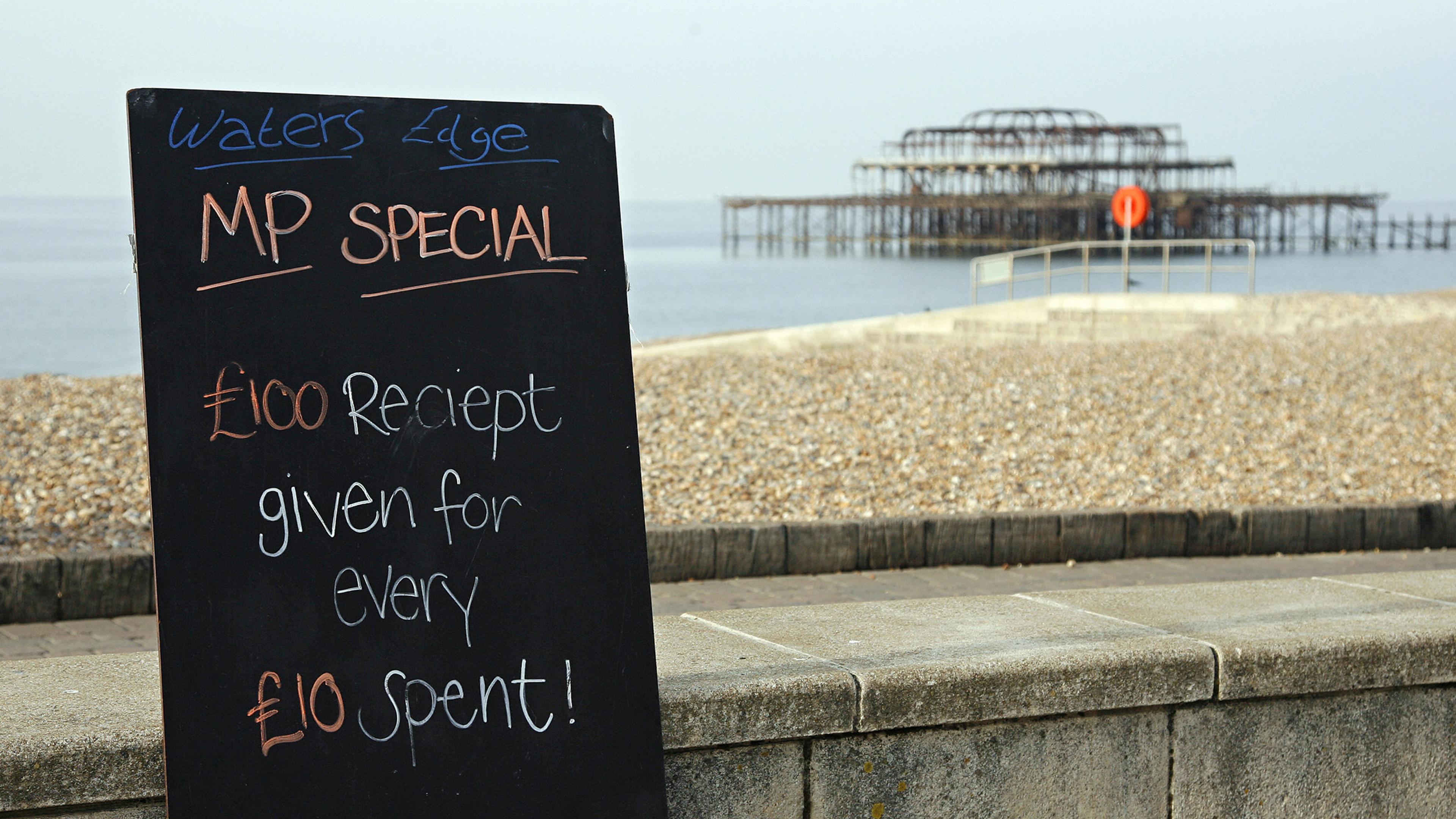Photo of a beachside blackboard offering a humorous deal near a weathered pier, stating “£100 receipt given for every £10 spent.”