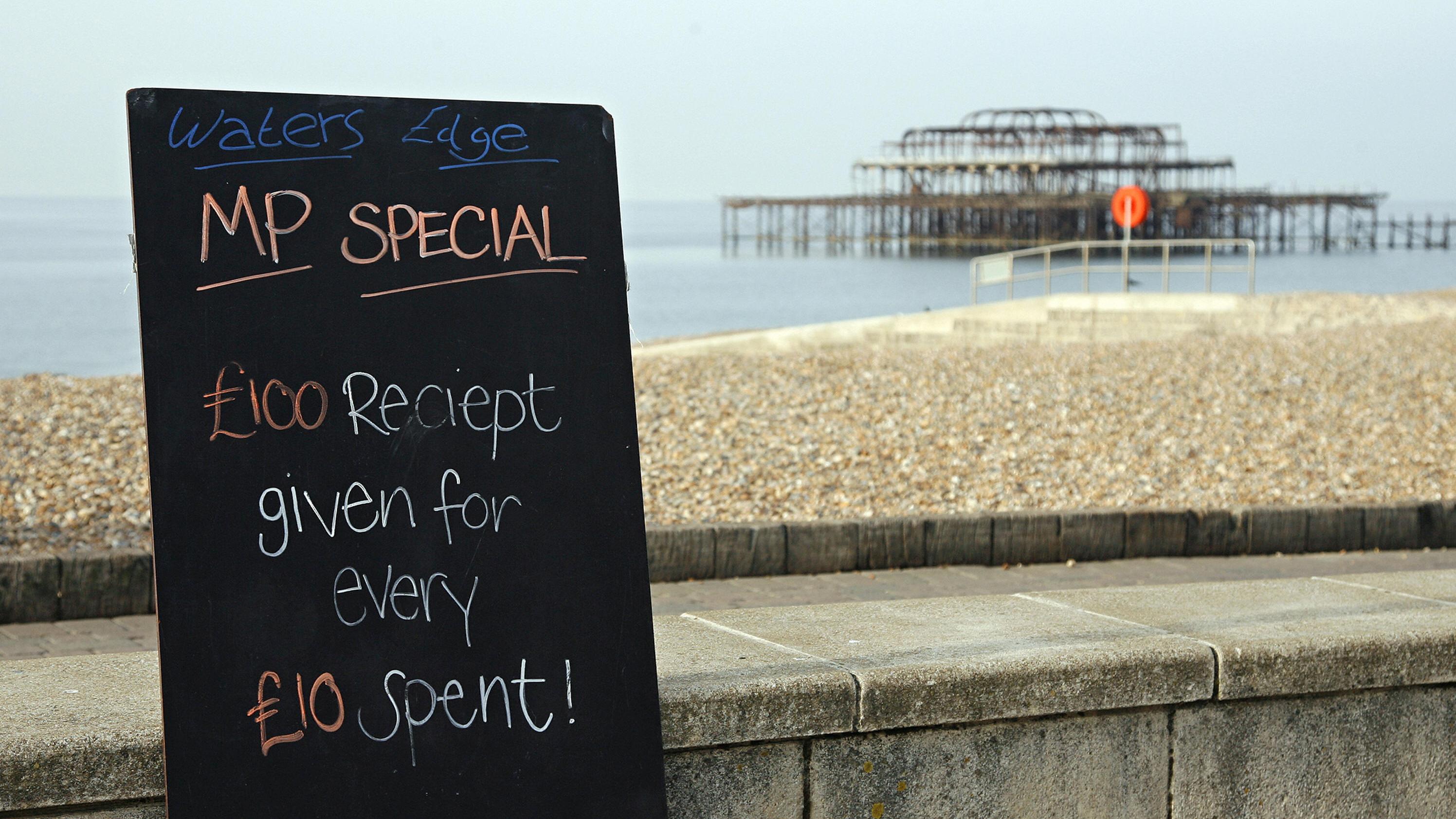 Photo of a beachside blackboard offering a humorous deal near a weathered pier, stating “£100 receipt given for every £10 spent.”