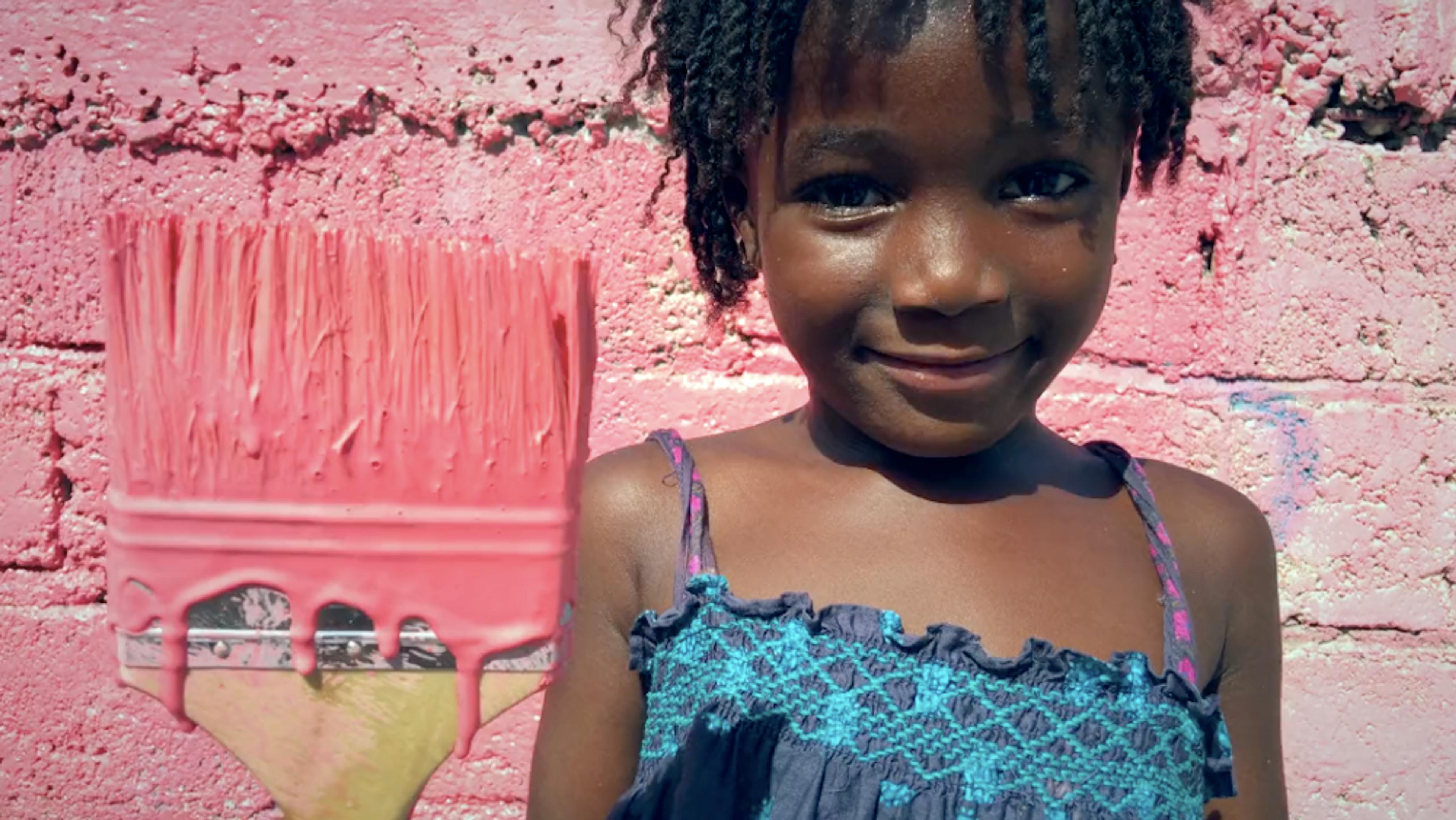 A smiling child in a blue dress holding a paintbrush with pink paint against a pink-painted brick wall.