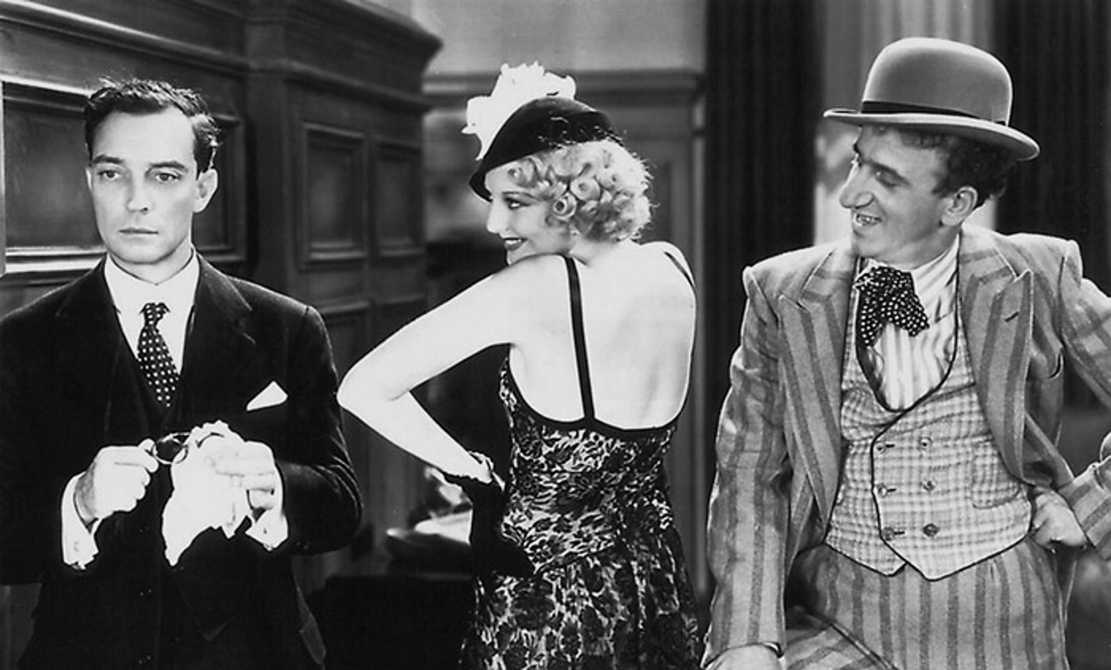 Black and white film still of three people in vintage attire. Buster Keaton looks serious while the woman and another man smile.