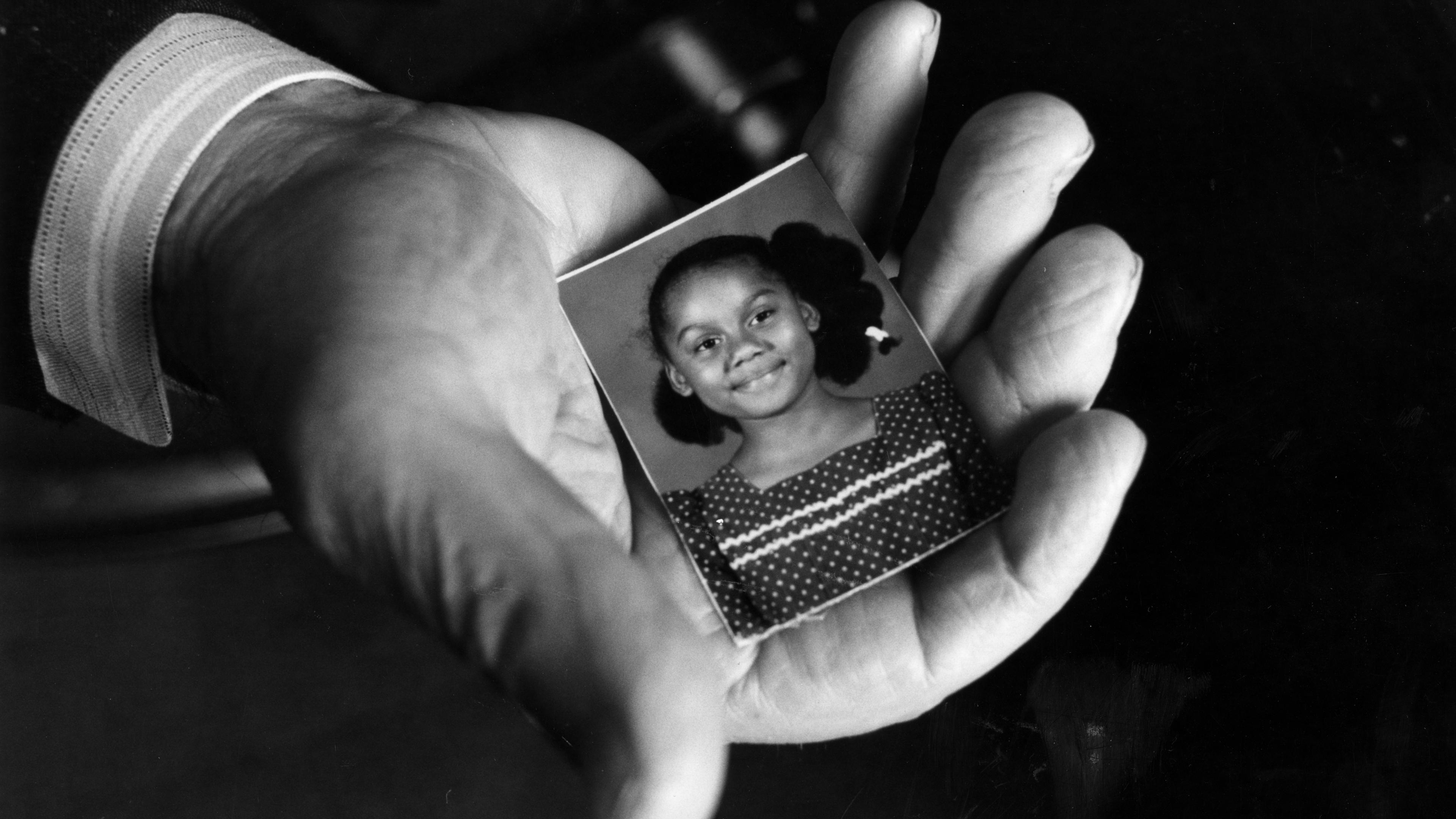 Black and white photo of a hand holding a small portrait of a smiling girl wearing a polka dot dress with braided hair.