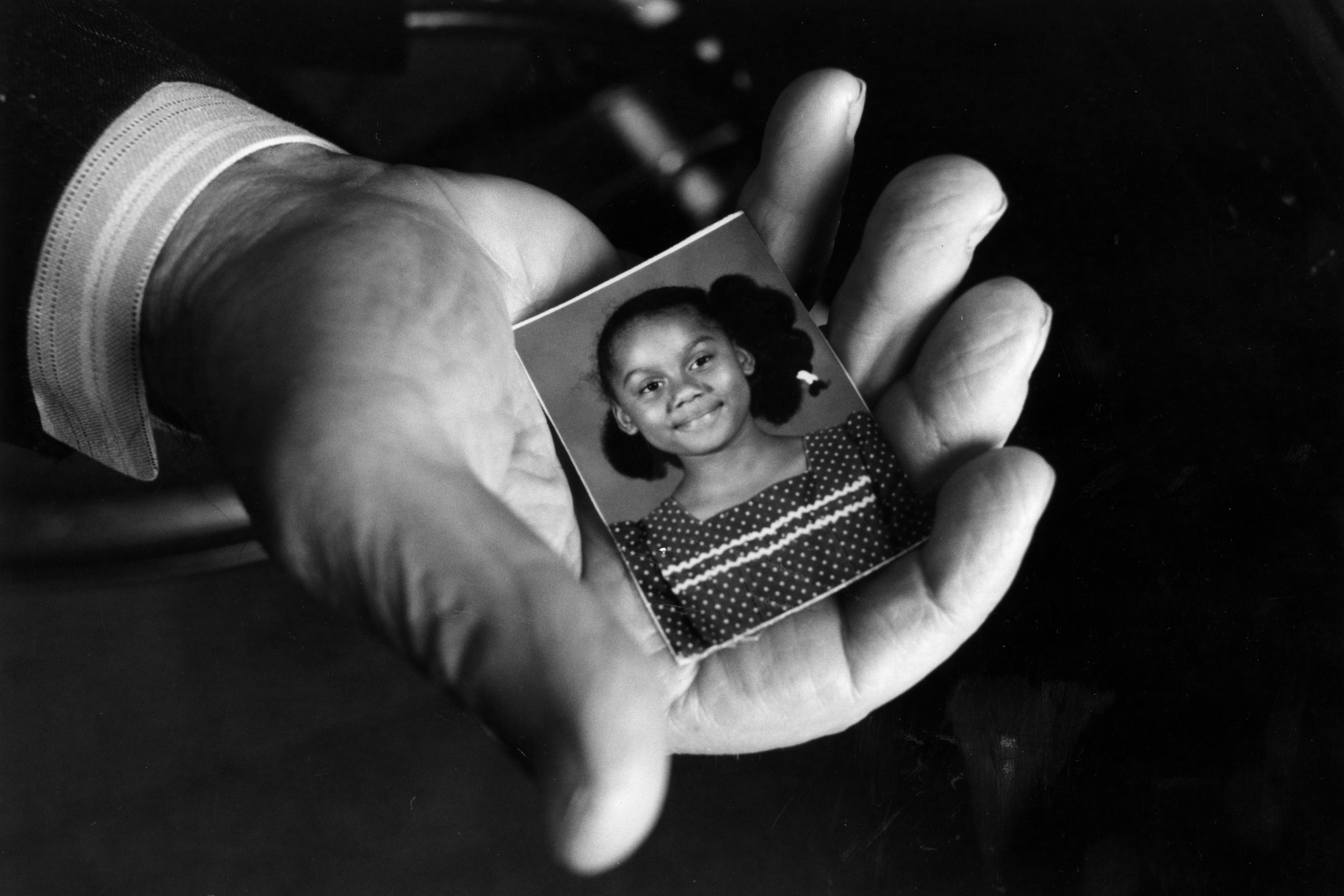 Black and white photo of a hand holding a small portrait of a smiling girl wearing a polka dot dress with braided hair.